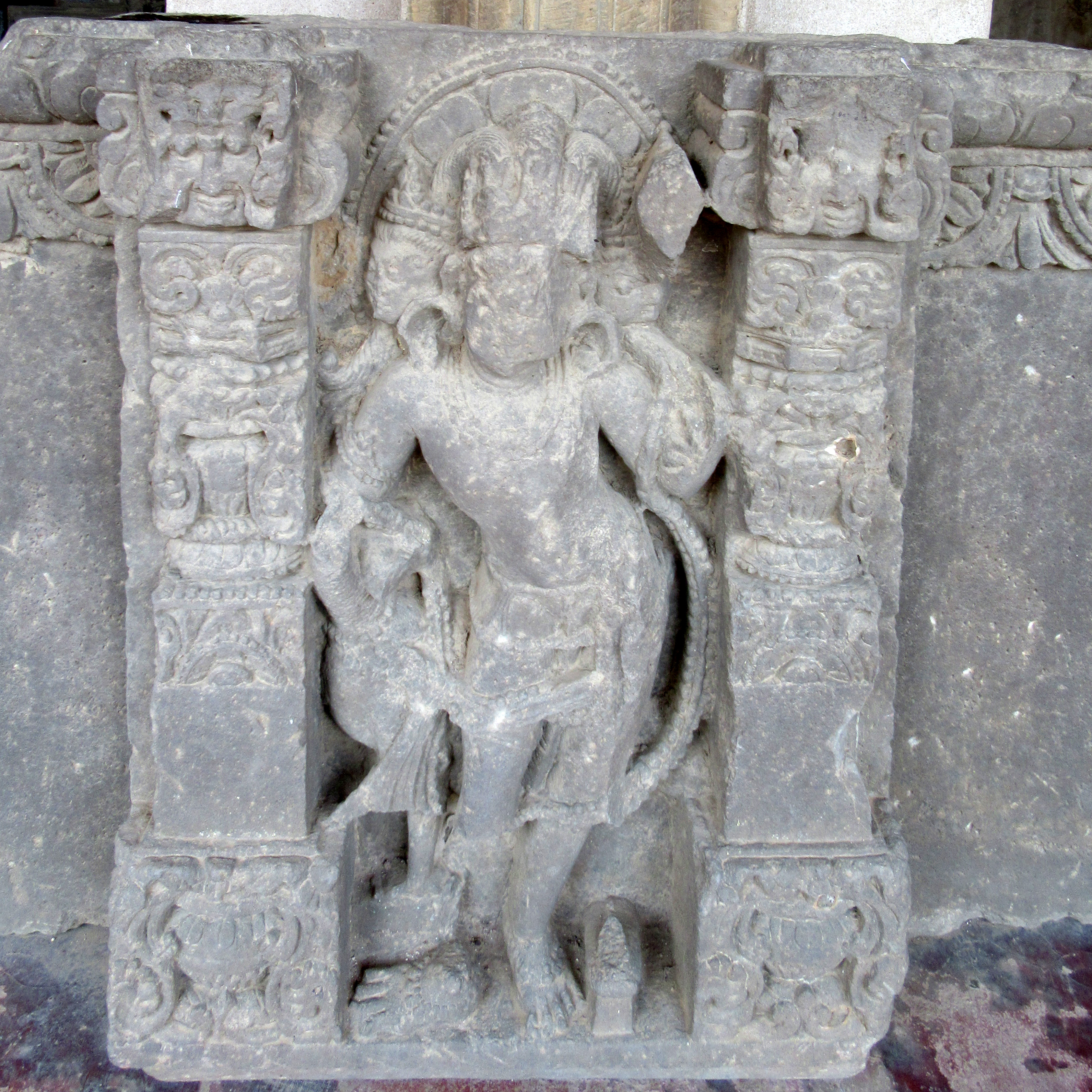 Image 19: A broken image of a two-armed male deity with three heads and faces. The central principal head wears a kakapaksha (a hairstyle with two side locks and a clearly visible middle section), while the others have small mukutas. He is adorned in a vanamala. The right leg displays a Vahana peacock. The halo is made up of lotus petals surrounded by pearls.