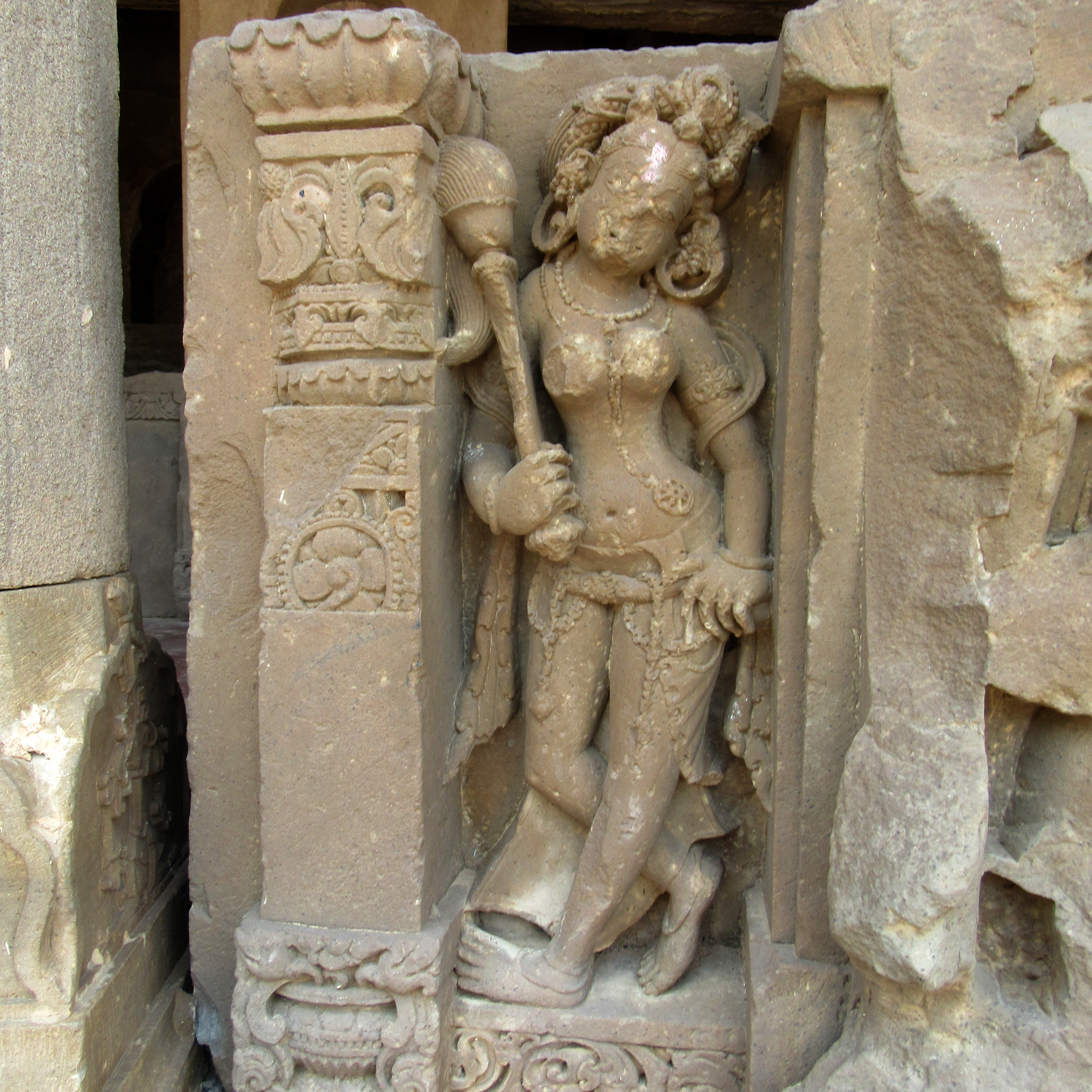 Image 20: A female attendant stands with her legs crossed. Her left hand is on her waist, while her right holds a fly-whisk. She has an elaborate hairstyle. The girdle that surrounds her waist has a hanging detail. A fine chain links it, and a leaf-like ornament hangs from it. This ornament, known as the lalantika, is recognised as an iconographic feature at the Harshatmata Temple.