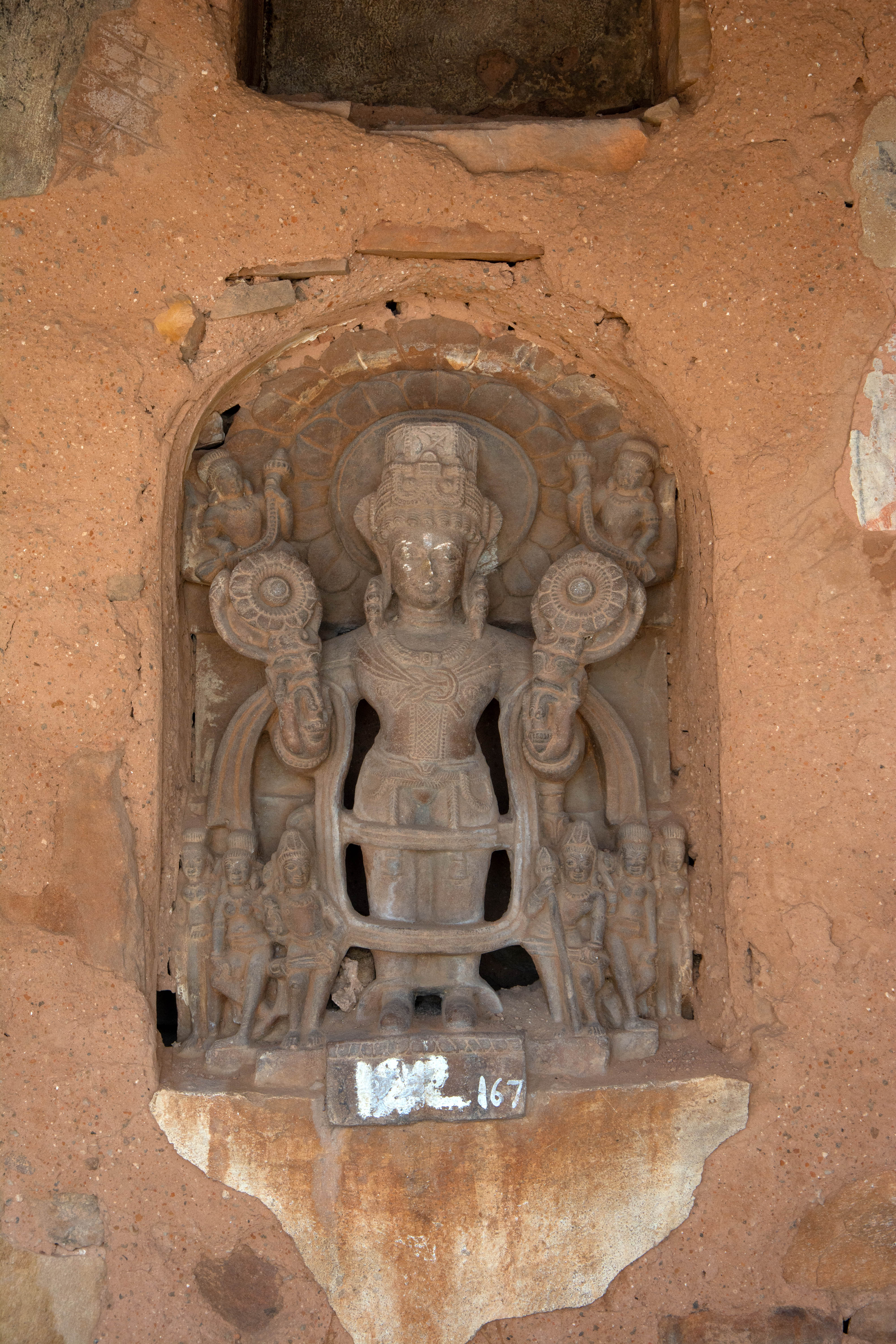 Image 3: This is arguably the oldest sculpture at the shrine, dating back to the 8th century CE, according to Ambika Dhaka. The sculpture significantly precedes the Harshanth Temple, built in the second half of the 10th century. The figure embodies the standard iconographic features of the Sun God, including fully bloomed lotuses in two hands, long tunics, boots, and retinue figures in pairs, representing his companions, Danda and Pingala, his wives, and his sons, the Ashvins,