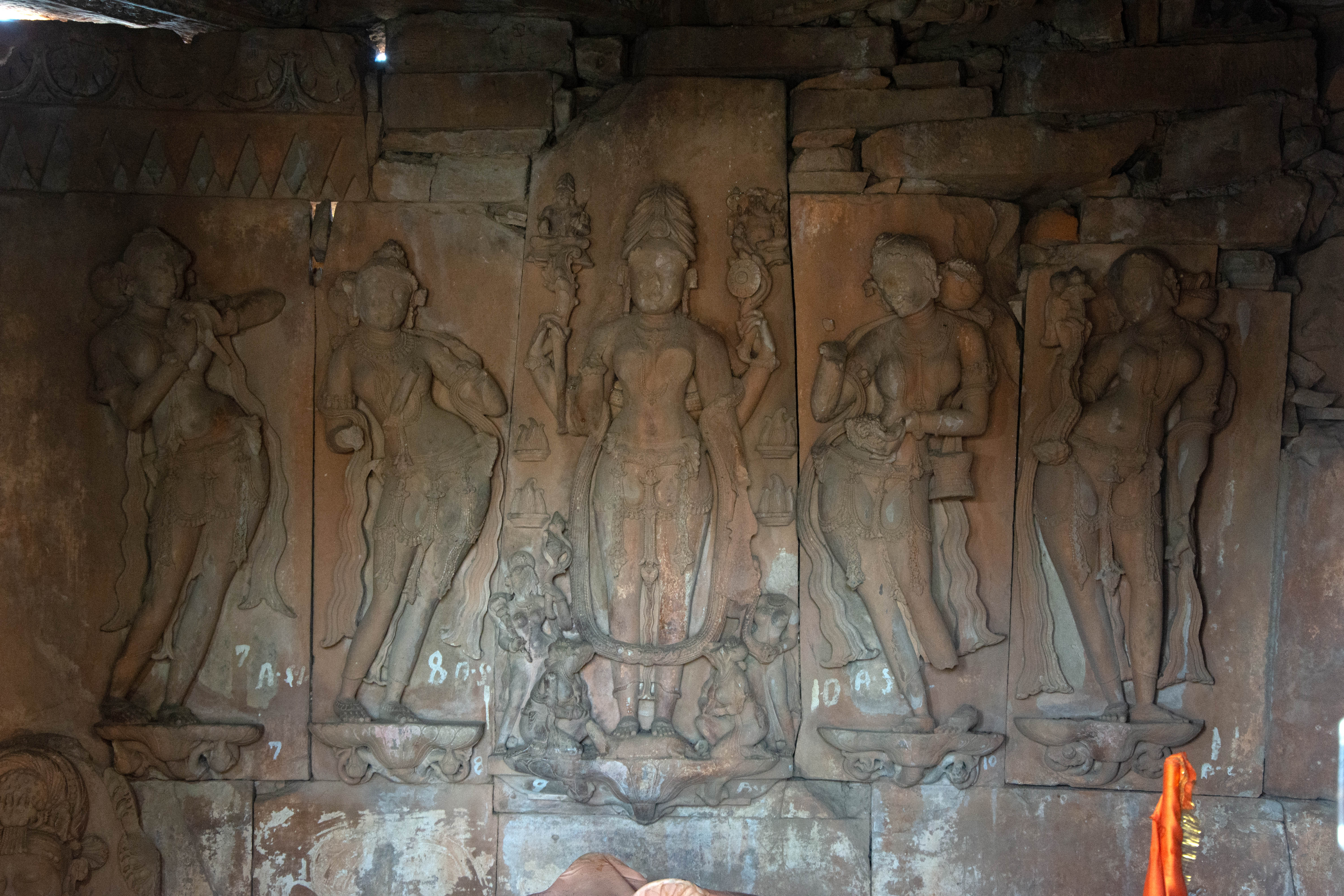 Image 10: Within the temple's sanctum, exquisite sculptures of nayikas adorn the walls, while a massive depiction of Parvati in penance, also known as the panchagni tapa, dominates the center of the principal/back wall. The image depicts a four-armed Parvati performing the panchagni tapa, with fires visible on either side. Known as Godhasana Gauri, she stands upright atop an iguana. On either side, there are two female attendants standing and two sitting.