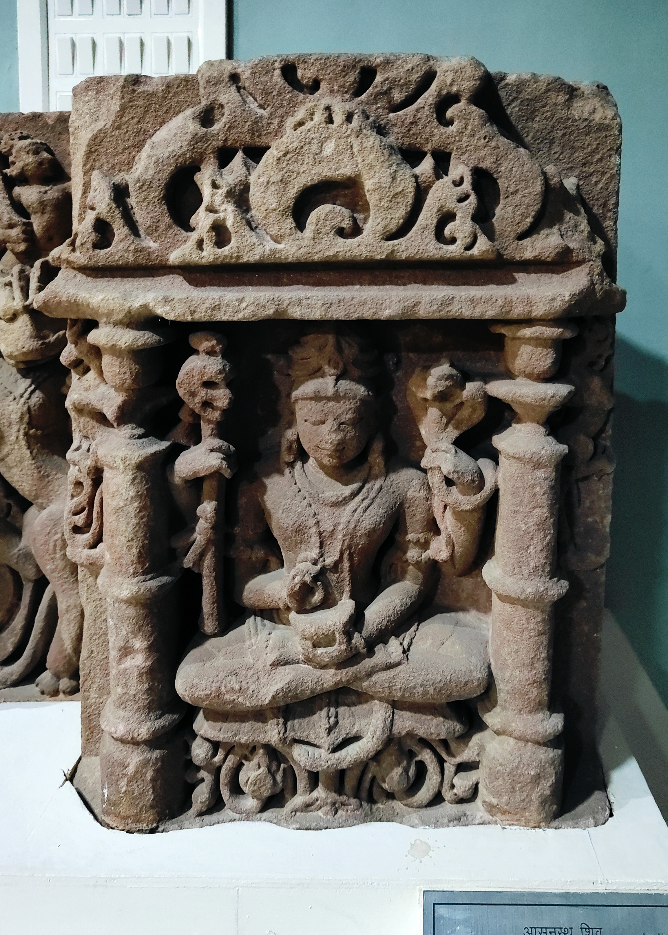 Image 15: Housed in the Government Museum, Sikar, this elegant Shaiva figure was part of a devakostha, or sculptural niche, on the walls of a now-lost Shaiva shrine. The figure has four hands, with the upper two holding a skull-sceptre and cobras. He holds a cup, or rather a skull, in his lower left hand, while the fingers of his right hand dip towards the potion inside. On the left is an image of Vyala, which was once placed in the temple wall's salilantara recess.