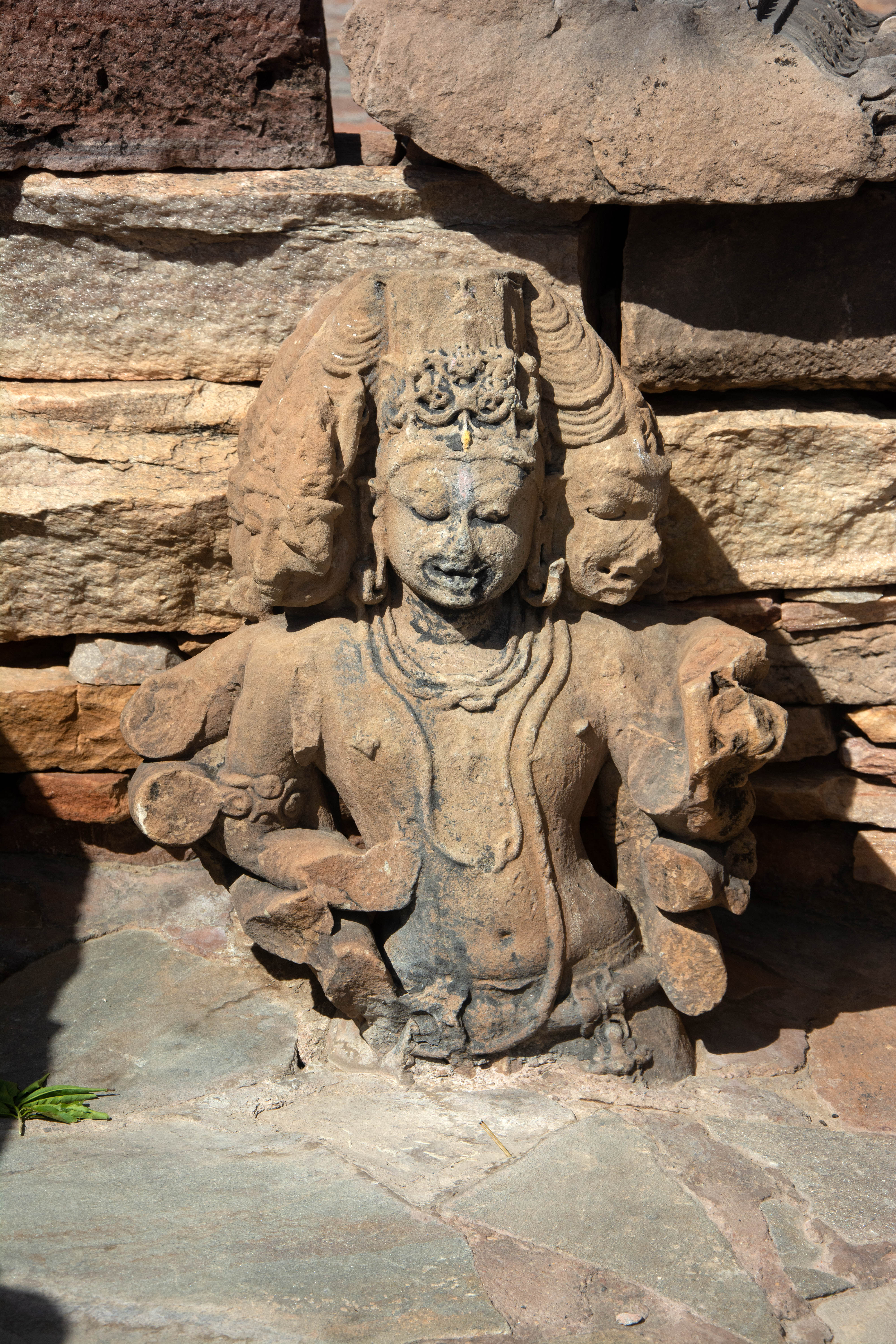 The depiction of this enigmatic deity, who appears to be Shaiva, features six heads and twelve hands. Although all hands are broken, one appears to hold an attribute in the form of a trident. The waist portion is buried in the earth. The headgear consists of a jatamukuta on the four side heads, typically associated with Shiva, and a kirita-mukuta on the front and back heads, usually associated with Vishnu.