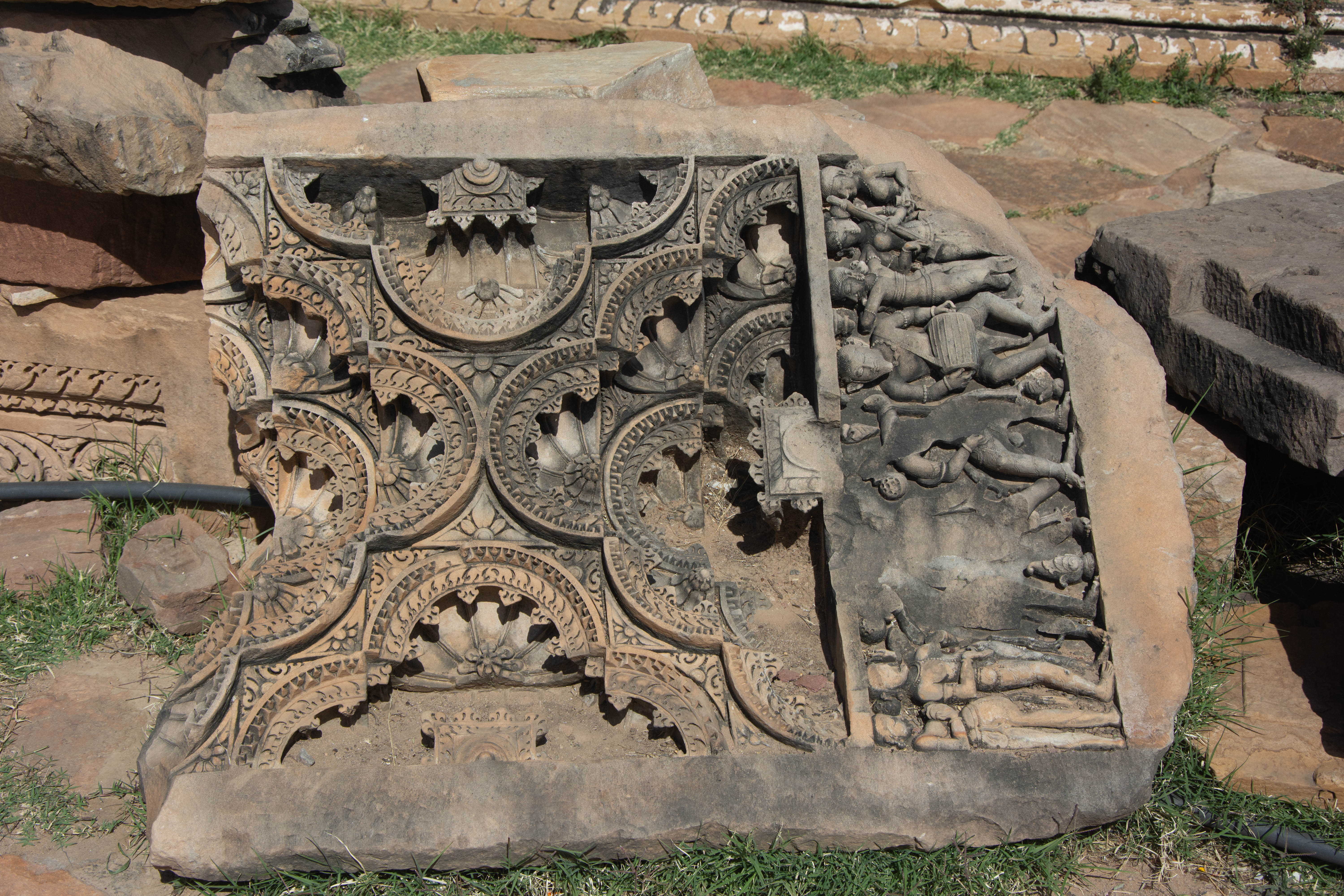 The elaborately carved temple ceiling features a panel on the right, carved with multiple male and female figures. A prominent bearded male figure is in the center, holding a drum. There is significant defacement in the central portion of the panel.