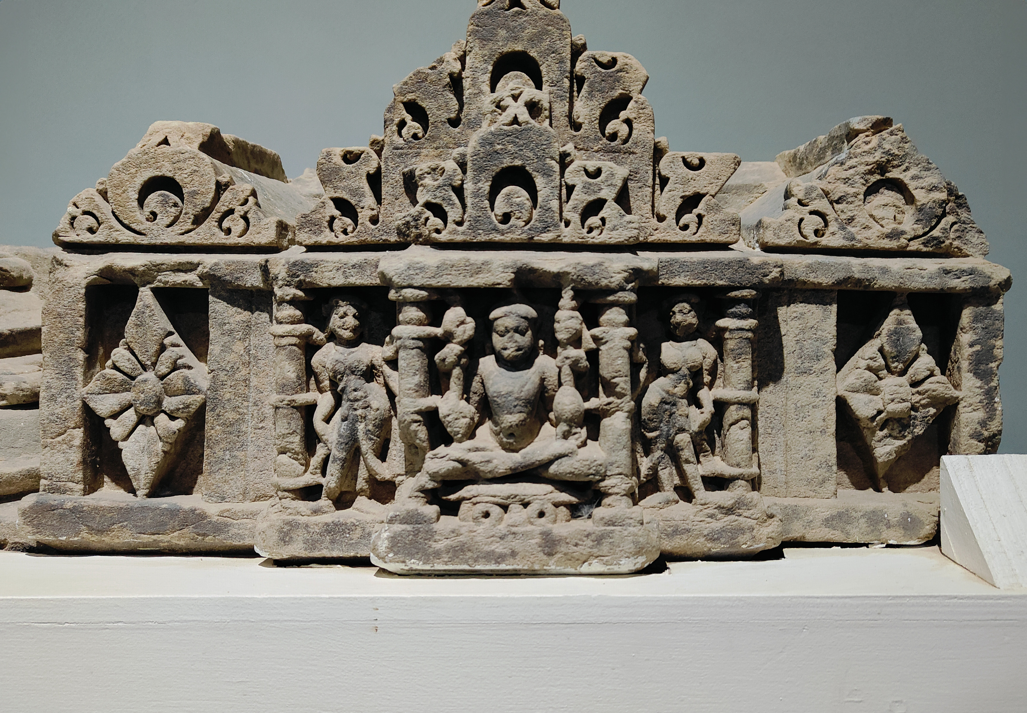 The four-armed seated figure, likely of Shaiva affiliation, occupies a central place in the architectural fragment. He holds a trident and likely a staff in his upper two hands, while the lower hands hold a citron in the left hand and an unclear object in the right. Despite the absence of a hyphalic representation, one could still interpret the figure as Lakulisha.