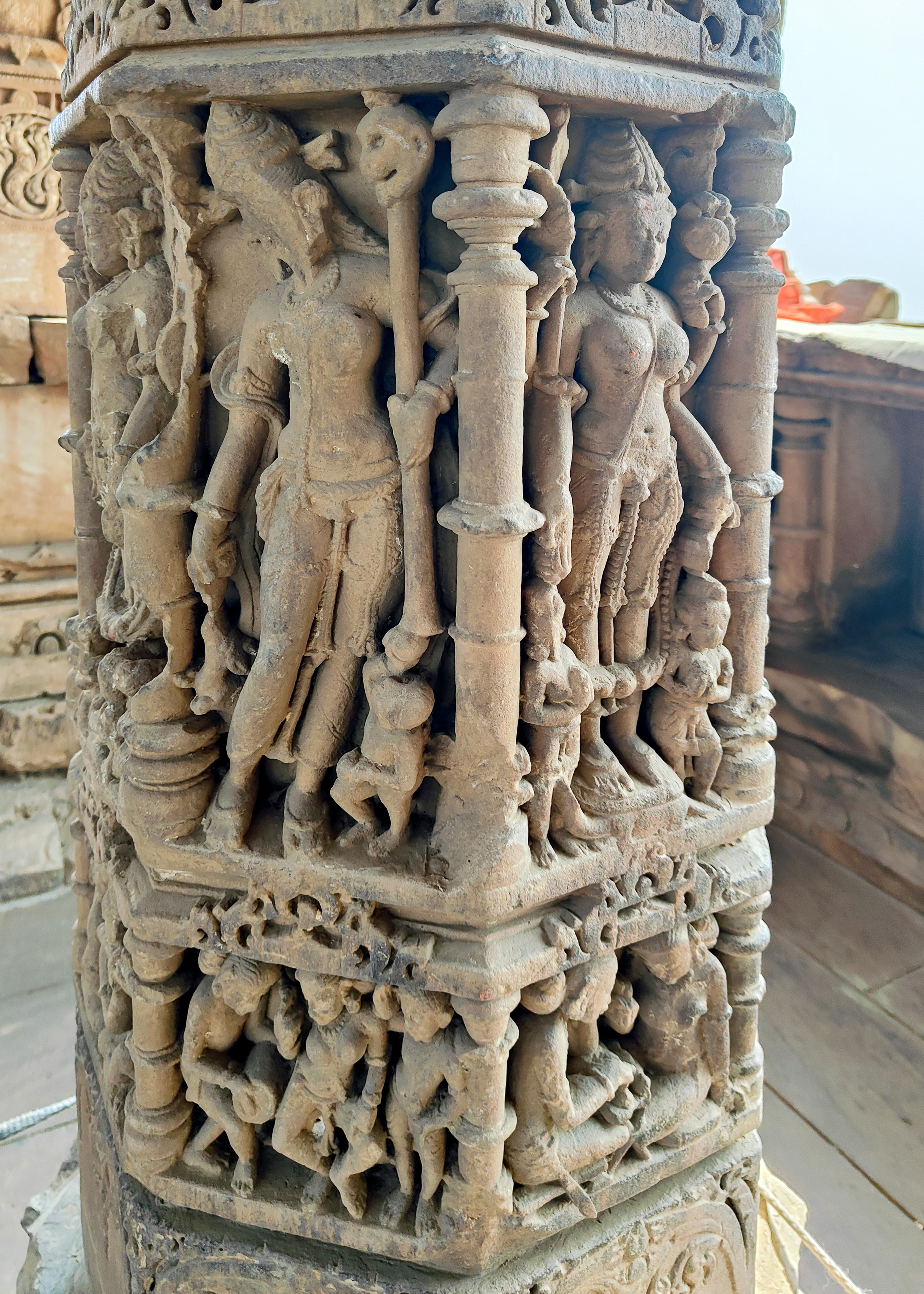 The four-armed goddess depicted here appears to be Goddess Bhairavi. In her right hand, she holds a staff crowned with a skull and an attribute resembling a mace. The left arm above is broken, and the lower one is placed near her vahana, a dog. The panel below depicts dancers and musicians.