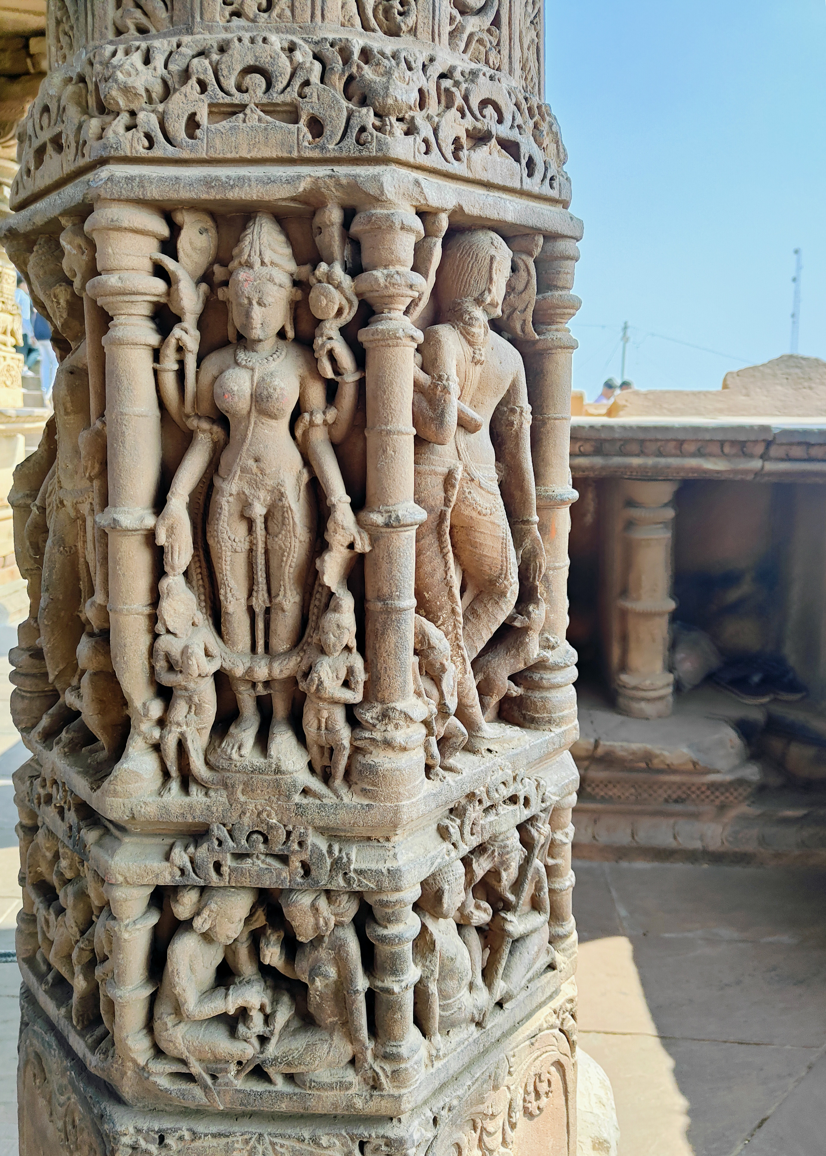 The four-armed goddess, likely Parvati, appears to be standing on a vahana that resembles an iguana. Two distinguishable features are a mala and a flower. The panel below depicts two figures engaged in a discussion over a text.