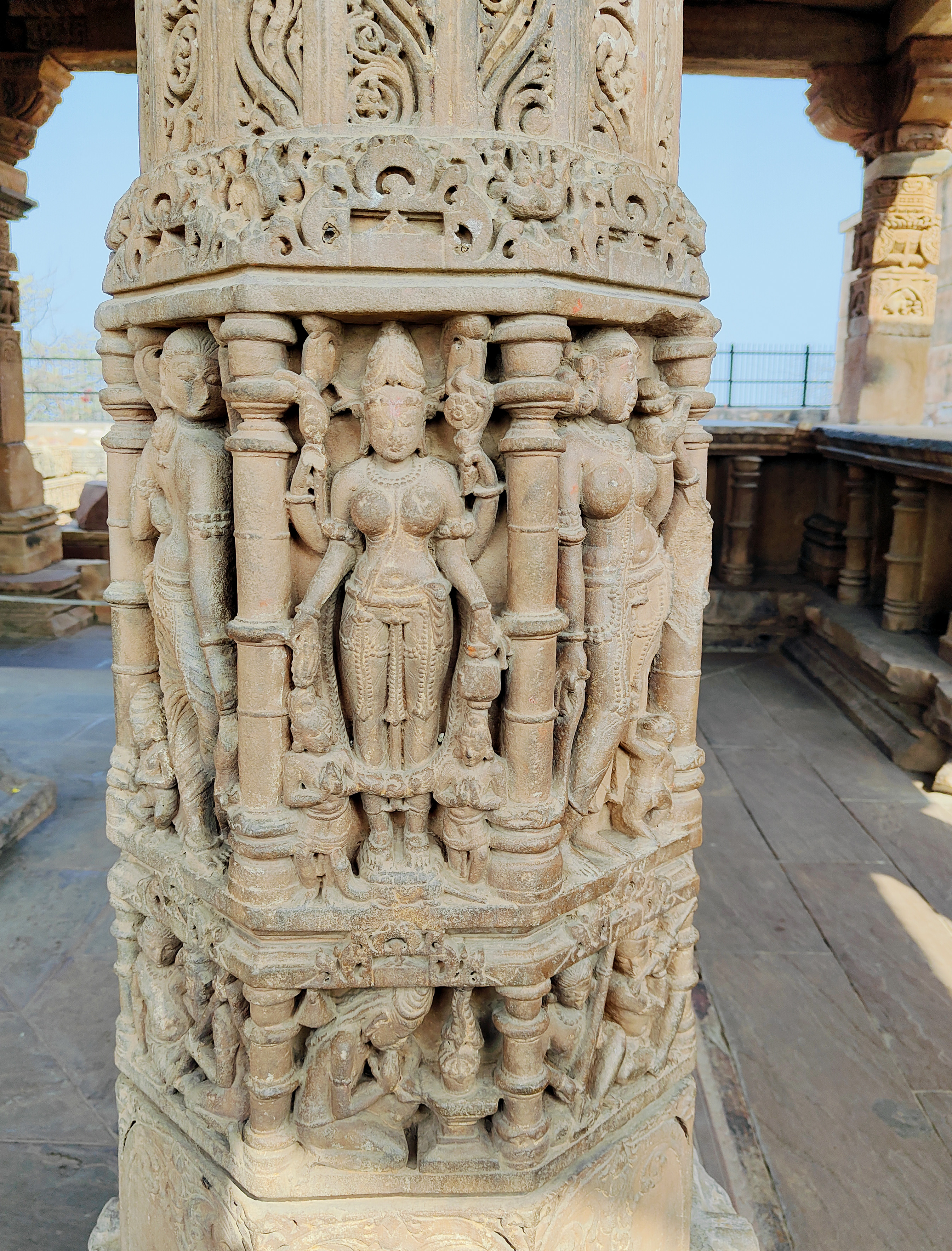 The depiction shows a four-armed Parvati holding a kamadalu and mala in her lower hands, and likely flowers in her upper two hands. On both sides, she is flanked by attendants. Below this scene is an ascetic with long matted hair, deeply adoring Shiva.