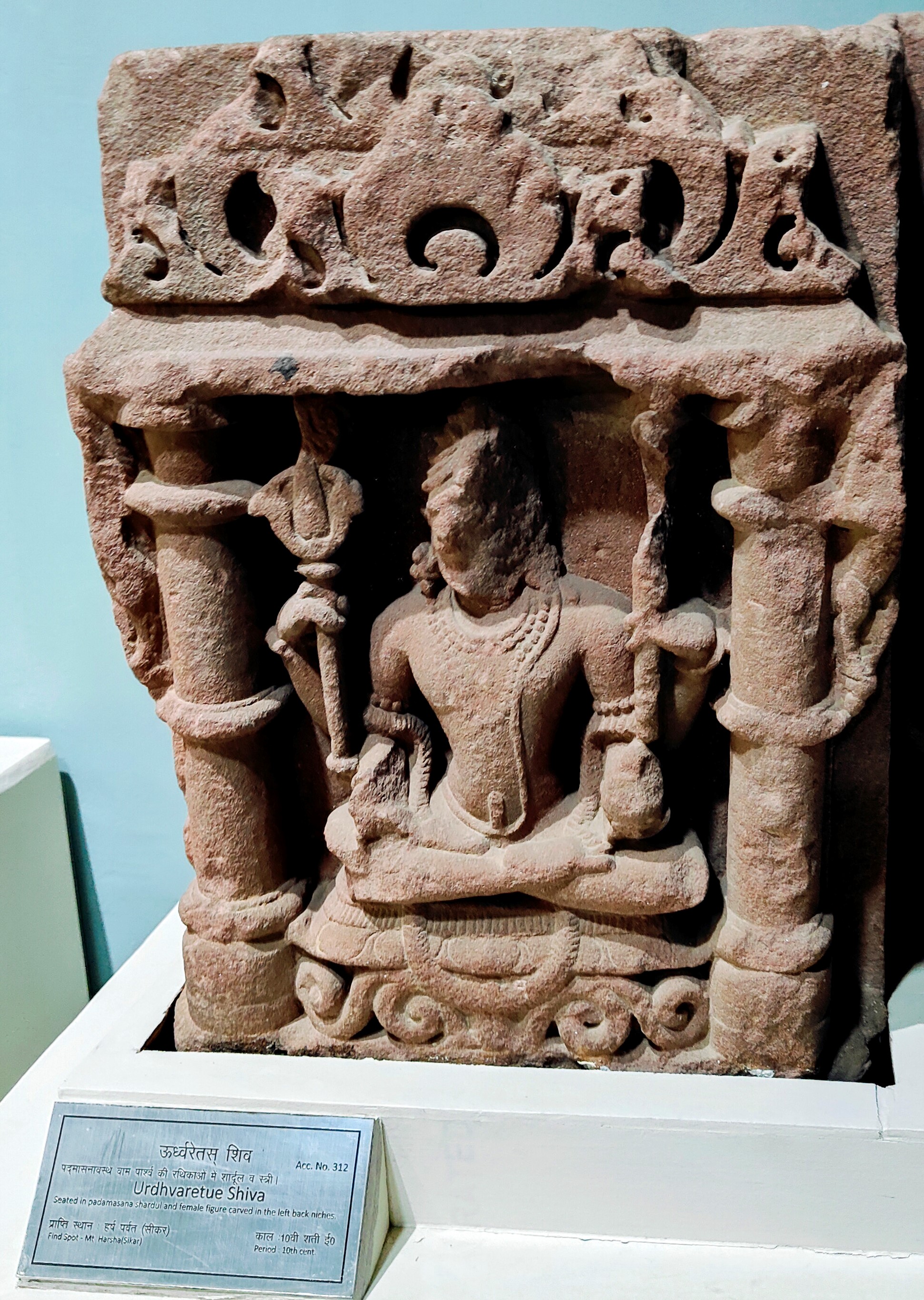 A now-lost Shaiva shrine may have included this Shaiva figure in a devakostha, or sculptural niche, on its walls. The Government Museum in Sikar currently houses it. The upper two hands of the four-armed seated figure hold a trident and a staff, while the lower two hands bear significant damage. The figure is ithyphallic, symbolising the Urdhvareta aspect of Shiva, indicating mastery over the vital energies through ascetic vigour.