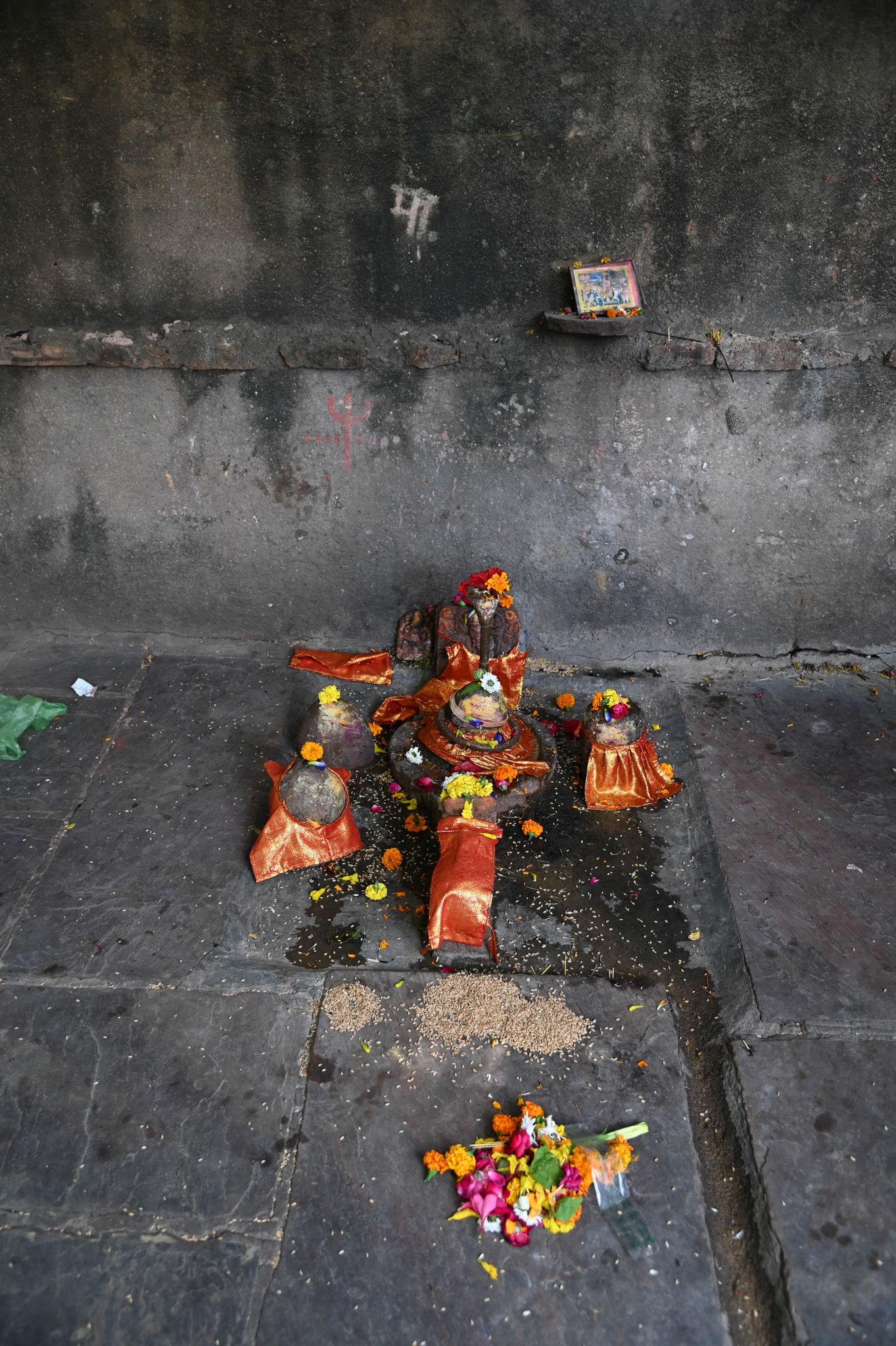 The stone platform on the left side of the stairs leading to the temple has a group of five sculptures. A Nandi sculpture precedes the central Shiva linga, adorned with a metal naga image.