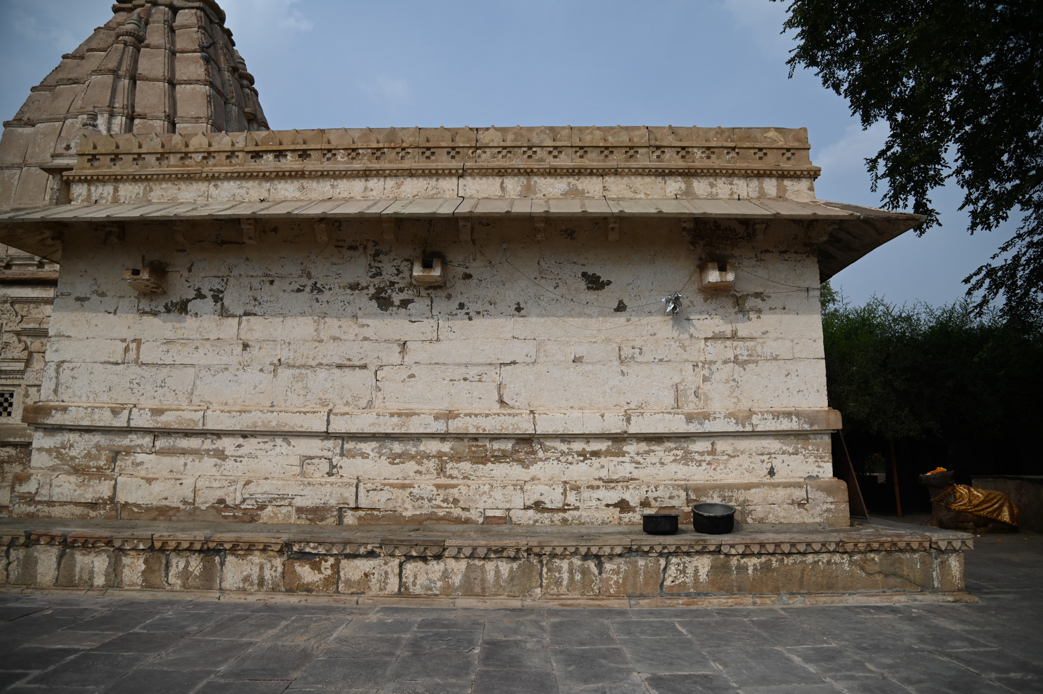 The temple's mandapa is a rectangular hall with simple, plain outer walls. It has a flat roof. There are no decorative elements on the walls, except for the flanking chhajja supporting the roof.