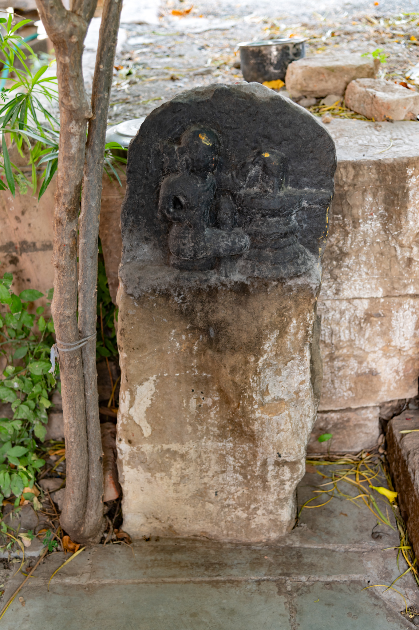Near the foundation of the subsidiary shrine (II), in the left side corner near the entrance of the Mahakal shrine, lies a stone stele. The stele portrays a Shiva Linga and a seated devotee who is worshipping the Linga. The Linga is held on a pedestal called yoni bhaga. The surface of the stele is quite weathered, so the details are not clear.