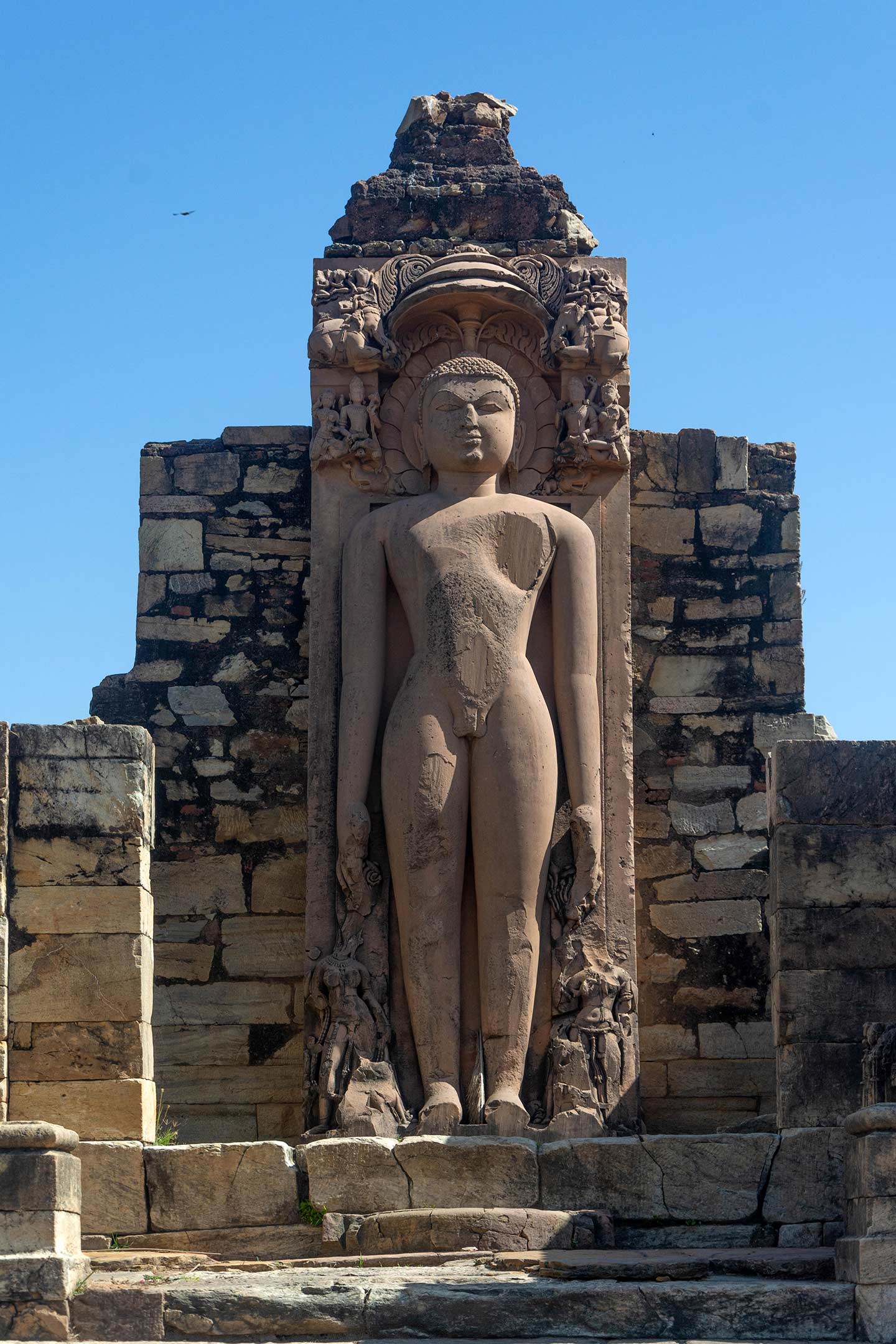 Image 2: The tall, nude statue of Jina Shantinatha stands in meditation posture with a parasol above it, placed at the back of the temple's inner sanctum. The statue has a calm face with a slight smile and curly hair, although some parts like the hands, feet, and chest are slightly damaged. Two female attendant figures near its feet are also damaged, making it hard to identify them. The sculpture is carved out of sandstone.