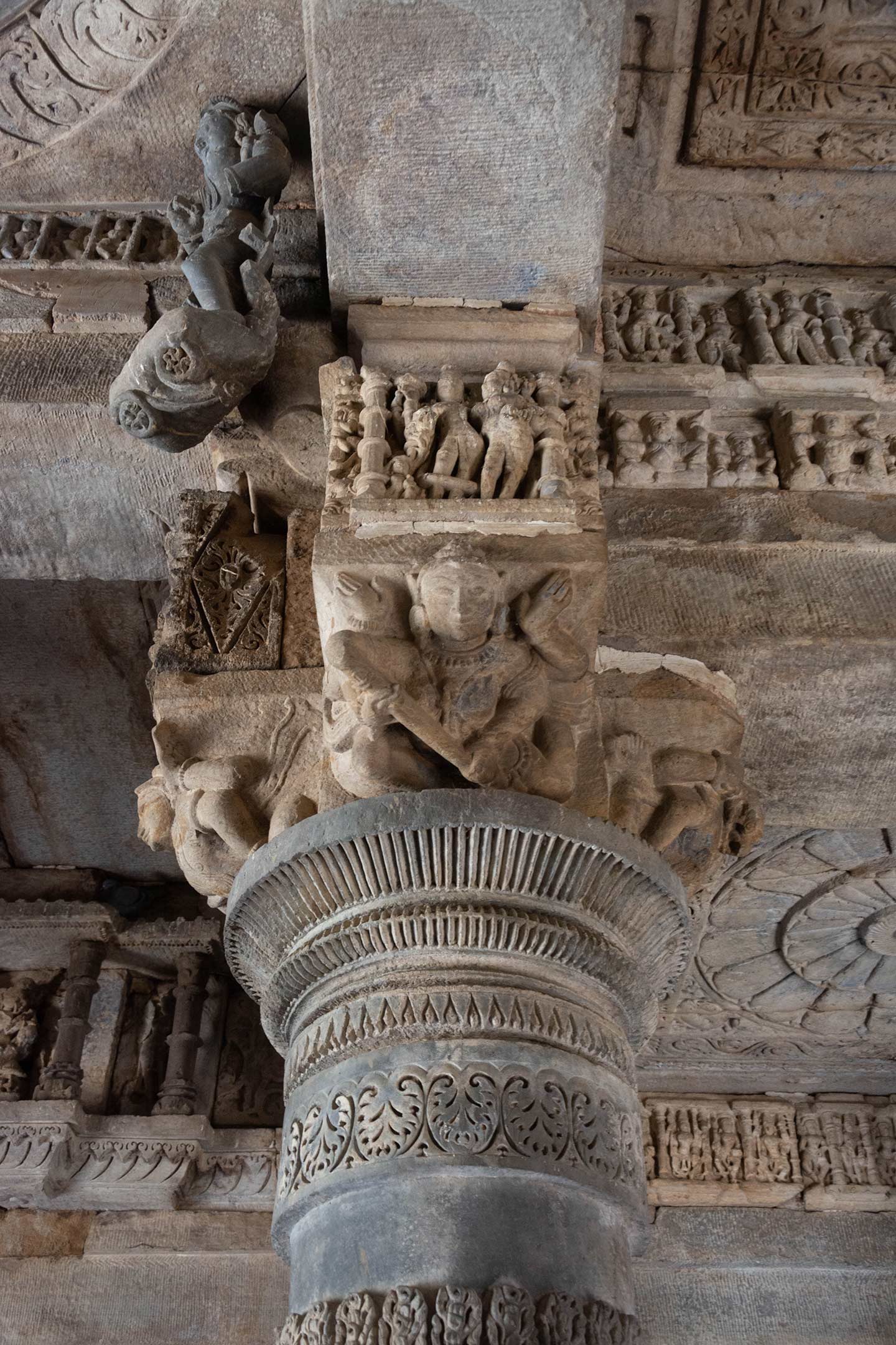 Seen here are the details of the pillar capital, the lintel above the pillar, and the ceiling base in the mandapa (pillared hall) of the Neelkanth Mahadev Temple. A four-sided bracket tops the large pot-shaped moulding of the pillar. Above these brackets, there are bharavahaka (load bearers) figures and small square sculptural panels. The frame structure's lintel beams feature friezes depicting fighting scenes and deity panels.