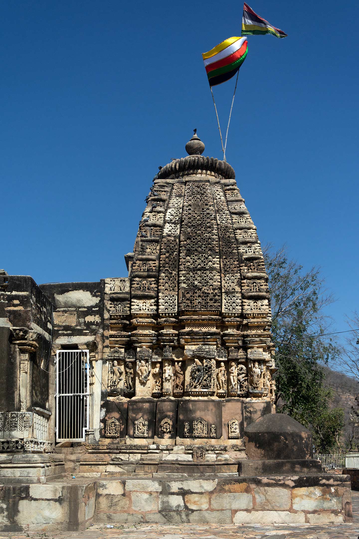 This is a view of the south-facing side of the shikhara (superstructure) central shrine at the Neelkanth Mahadev Temple. Since the central shrine has undergone renovations, the plinth, wall, and spire portions are in place. In plan, the temple is a variety of Latina nagara (mono-spired) shikhara and pancharatha (five vertical offsets or projections on each side). The kumbha (pot) moulding at the plinth level has undergone renovation. They may have replastered the deities on the kumbha's faces during this process.