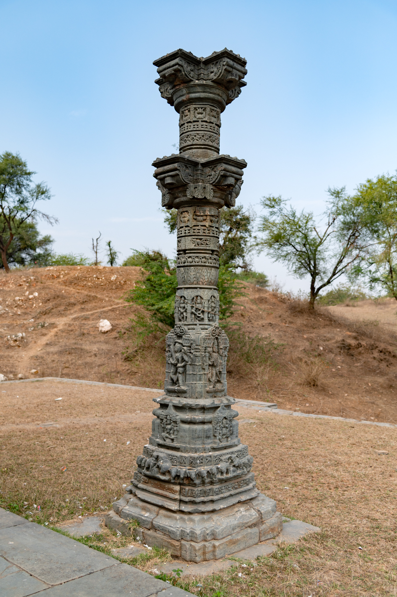 The existing pillar likely originated from a torana (decorative arch), which was a component of the archway on the northern side of the temple. The intricate carving of the pillar is based on a multi-moulding plinth, the base of which features a row of elephants known as the gajathara (elephant band). The lower shaft of the pillar has three sets of sculptures, and the topmost part has two brackets. Attaching the torana to the bracket that remains on the pillar is essential.