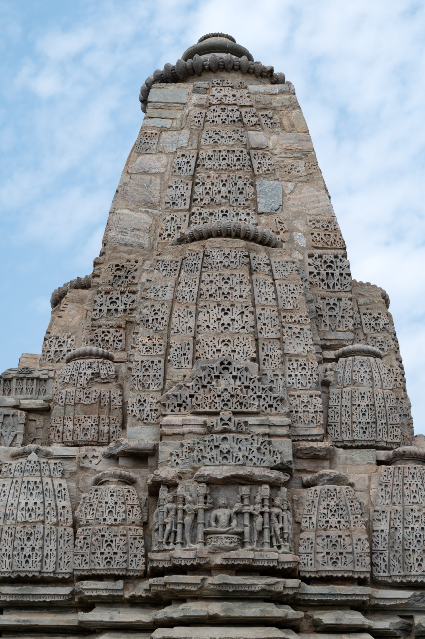 The Shiva Temple's shikhara (superstructure) can be seen up close. The Shiva Temple features the shekhari (multi-spire) variety of shikhara, where miniature shikharas and half portions emanate from the central spire. All the small and big shikhara aedicules are designed like individual Latina shikhara (mono-spired) with gavaksha (dormer windows) designs. The renovated portions of the shikhara were remade in plain stone blocks. The shikhara's base features an image of a seated Lakulisha, depicted nude, holding a staff, and flanked by two attendants.