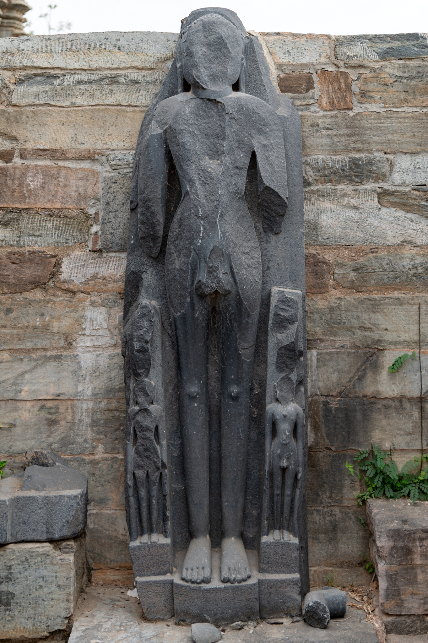 This is a colossal sculpture of a Jain Tirthankara standing in kayotsarga (dismissing the body) posture, found in the ruined shrine to the west of the central temple. The inscriptions at the base of the free-standing statues date from the 16th century CE. Black stone, primarily granite, forms the sculpture's carving.