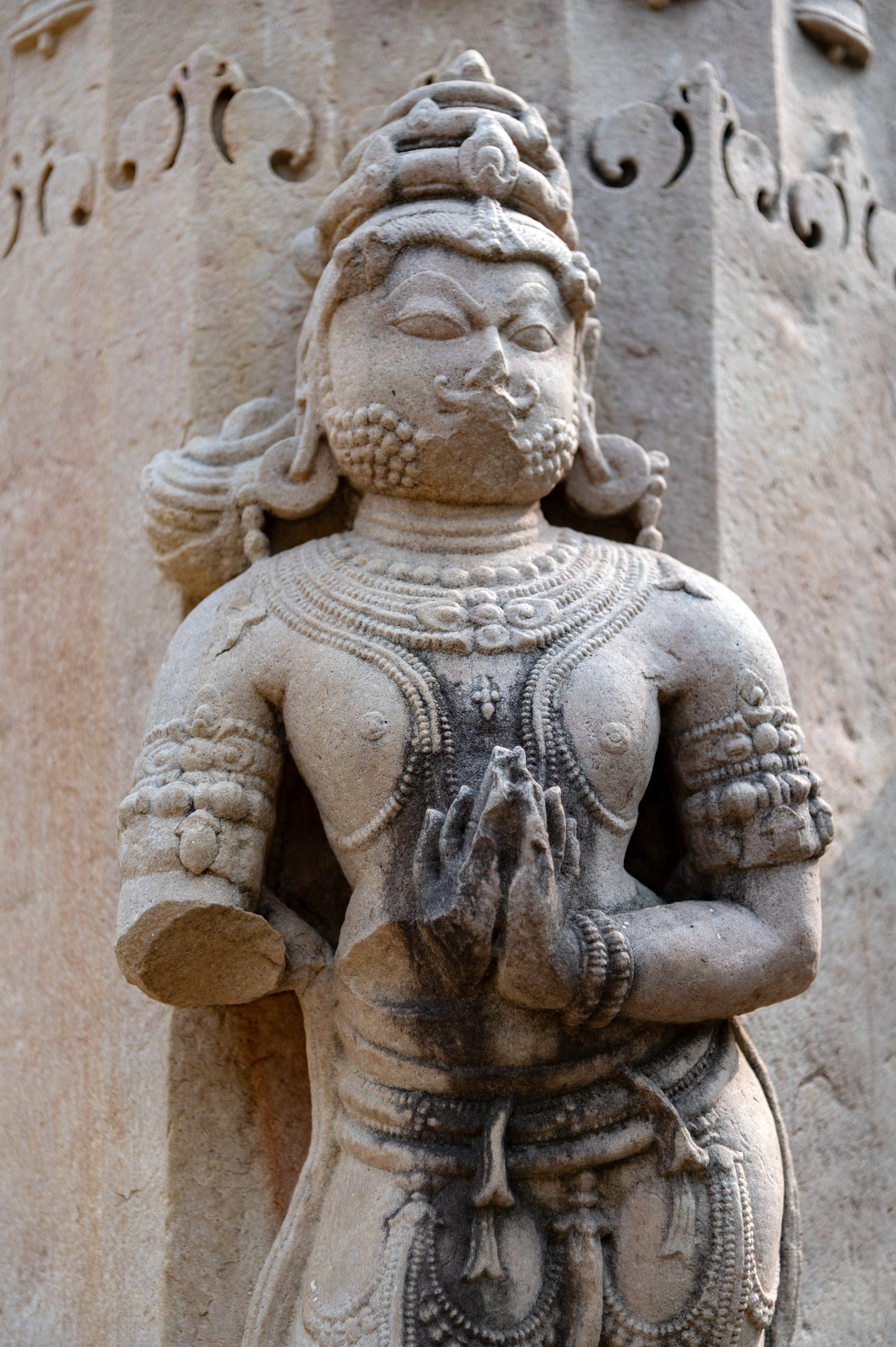 The male figure on one of the pillars of the torana (ornamental gateway) depicts a standing male figure standing with folded hands near his chest. This figure is likely a deity as he wears a crown and heavy jewellery and has a mark on his chest commonly found on Jaina deities. His charming visage features raised eyebrows, a mustache, and a beard.