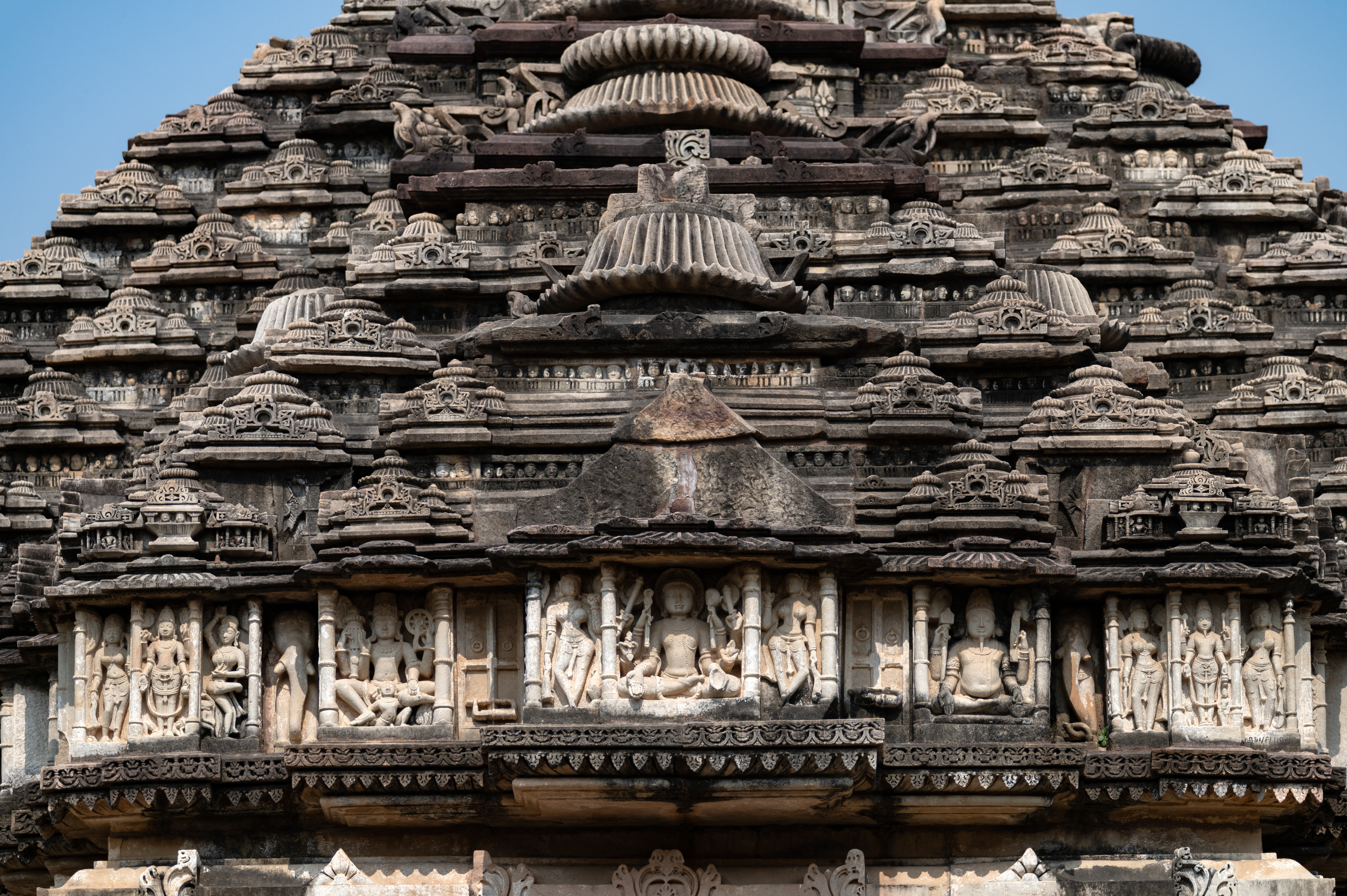 The west pediment on the shikhara (superstructure) of the separate rangamandapa (an open type of pillared hall used for ceremonial rituals) depicts the Trinity. At the centre, Shiva is shown in his Bhairava form, flanked by Vishnu on his right and Brahma on his left side. There are two other gods, one on each end. The Trinity is shown seated while the other two gods are shown standing. A six-armed Bhairava is shown holding his multiple attributes, and his aggressive aspect is demonstrated through his fangs. A four-armed Vishnu is shown with his attributes, seated on Garuda, and Bramha is also shown seated, holding his attributes. The other two gods are also four-armed and hold their respective attributes.