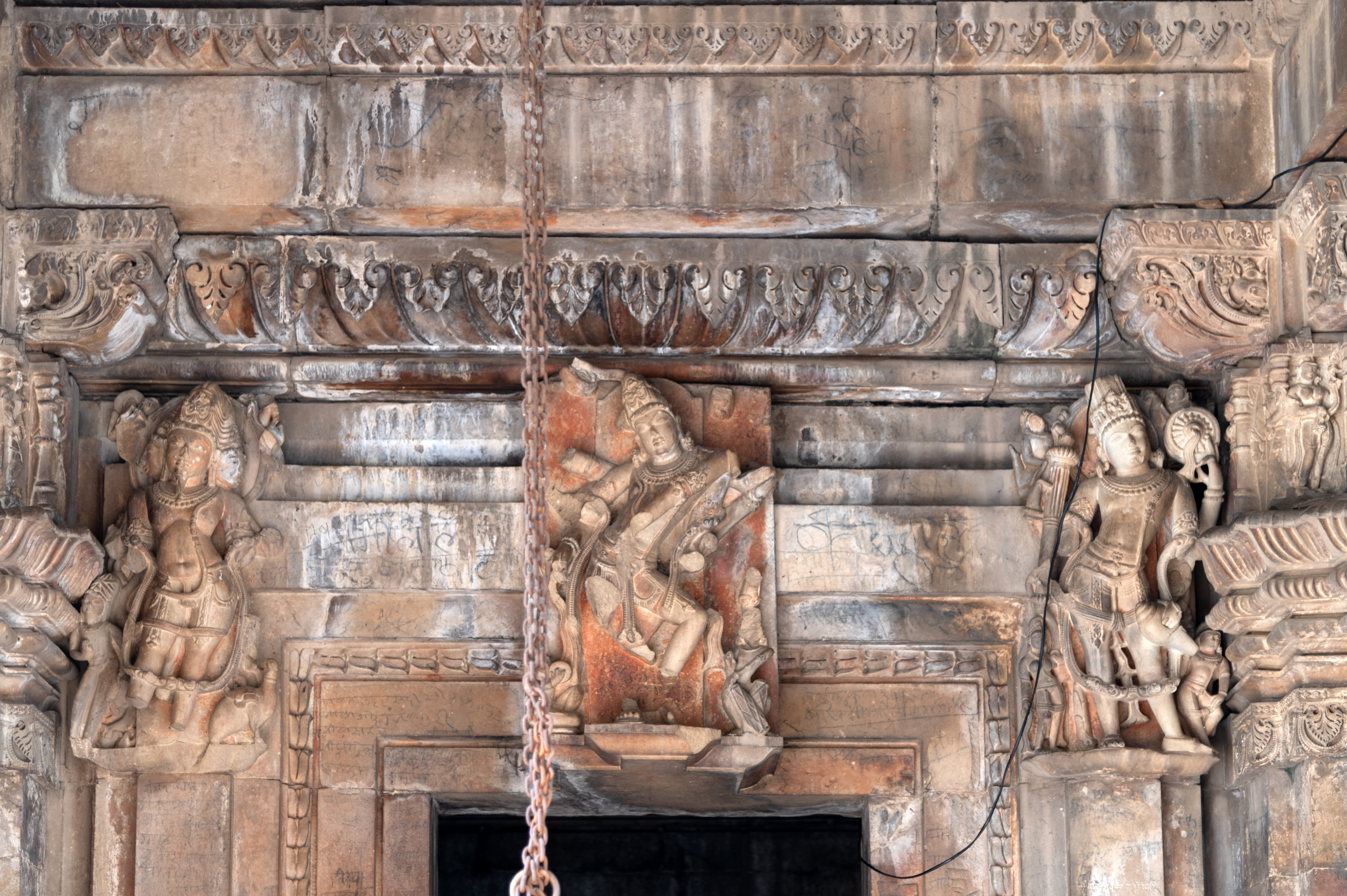 The lalatbimba (lintel) of the doorframe is relatively plain, featuring three large panels placed at the centre and two on the extreme ends depicting the trinity—Shiva, Vishnu, and Brahma. In the central panel, an ethereal sculpture of Shiva in the form of Nataraja is carved. To the proper left, a sculpture of Vishnu is carved, while to the proper right, a sculpture of Brahma is represented. Nataraja had multiple arms, now all broken, with an elegant posture and glorious face drawing attention. He is accompanied by two musicians who are playing musical instruments. The charming figure of Vishnu is shown holding shankha (conch), gada (mace) and chakra (discuss). The three-faced Brahma is also carved with four arms, now broken, with his vahana (vehicle or mount) at the bottom.