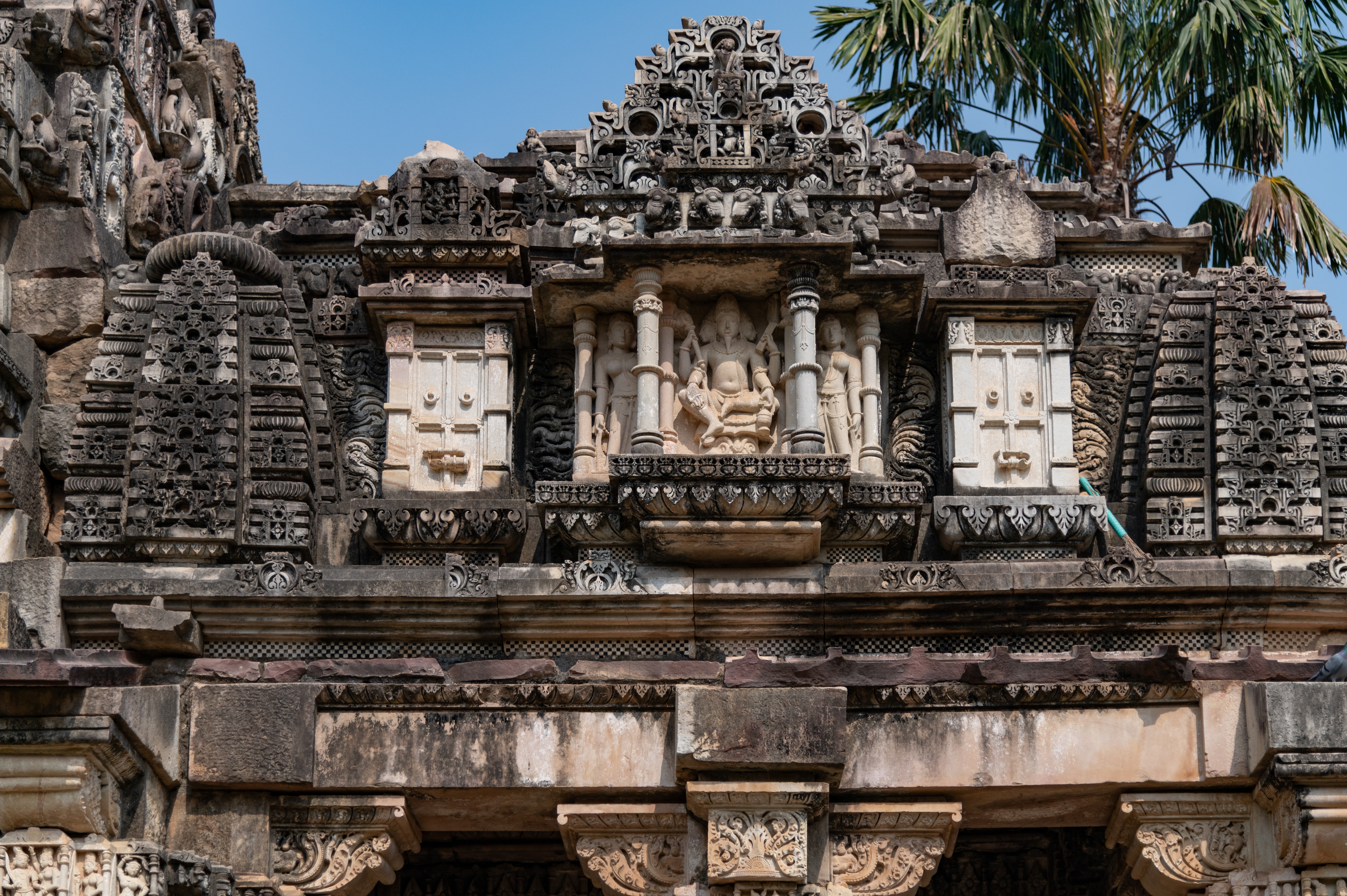 The sukanasa (antefix above the roof of the kapila) is placed above the antarala (vestibule or antechamber), which serves as the frontal pediment of the garbhagriha’s (sanctum sanctorum) shikhara (superstructure). The southern side of the sukanasa is carved with a distinct scene depicting a miniature balcony where three beautiful women are shown talking to each other. Above the balcony, an udgama (pediment of interconnected chaitya dormers) is placed, depicting a four-armed goddess seated on a lotus seat, flanked by two attendants.