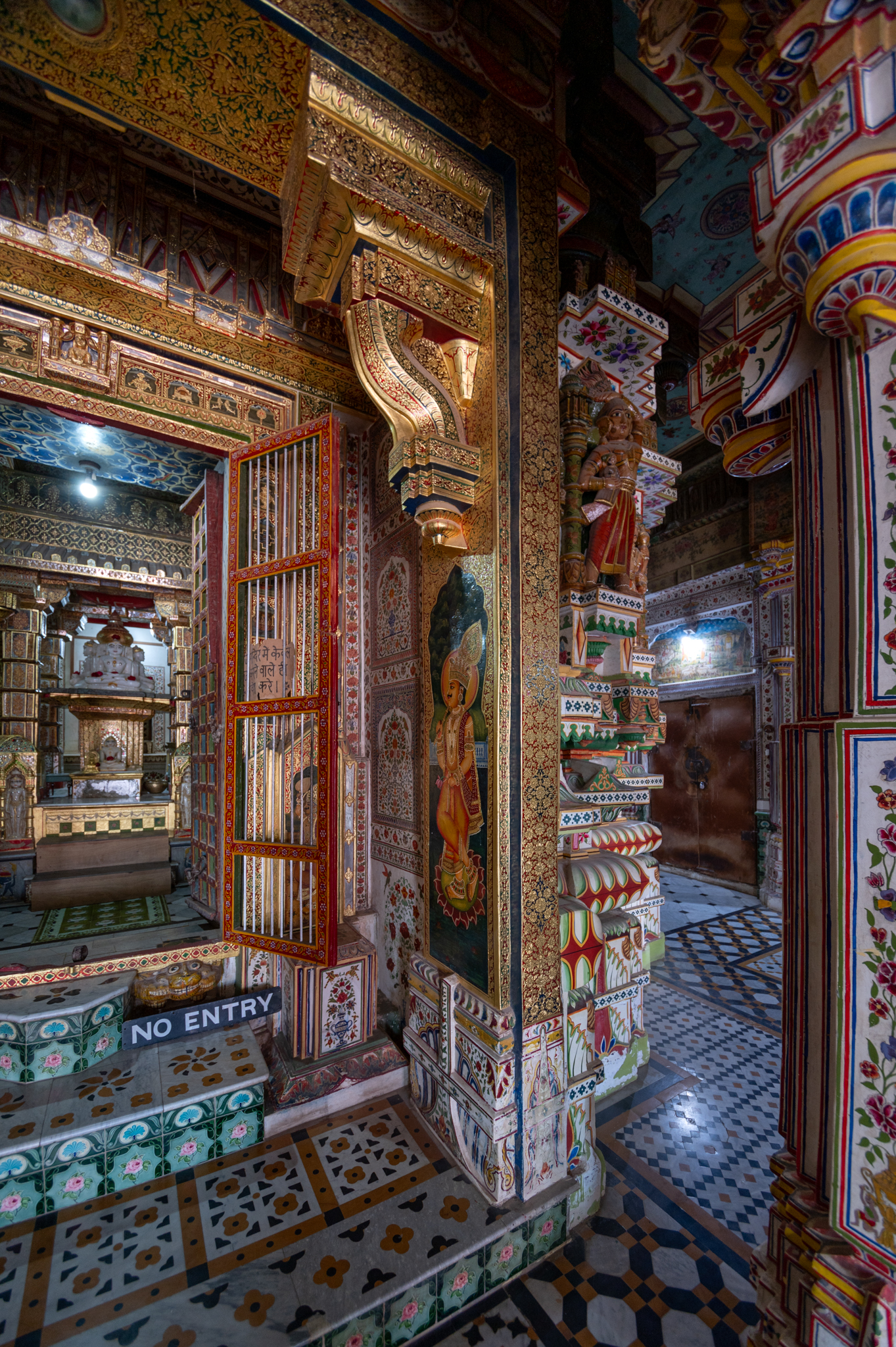 Seen here are the pilaster and bracket details of the garbhagriha (sanctum sanctorum) entrance. The inner face of the pilaster has a painting of a doorkeeper, and the original surface, probably in stone, is covered entirely in meenakari (enamel) goldwork. The lalatbimba (lintel) has a figure of Jain Tirthankara. The use of gold embossing on the doorway is remarkable. It sets apart the garbhagriha from other parts of the temple interior.