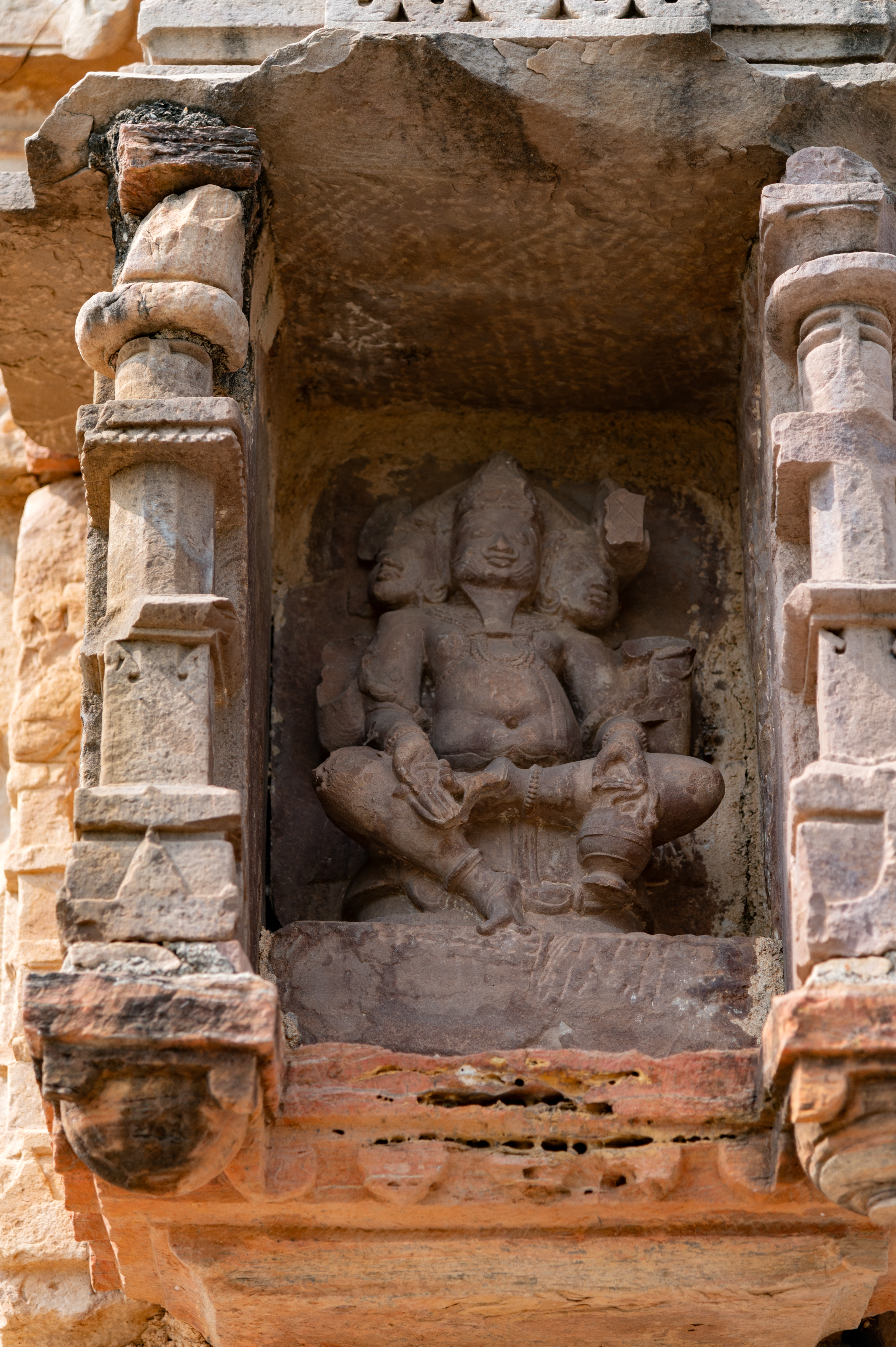 The bhadra (central offset) positioned on the rear (east) wall of the garbhagriha (sanctum sanctorum) of the Undeshwar Temple features an image of Brahma. The deity, albeit significantly damaged, is depicted seated on a pedestal. He is portrayed with three faces and four arms, although his upper arms are broken. In his lower left hand, he holds a kamandalu (water pot) while displaying the varada hasta (hand gesture of conferring a boon) with his lower right hand. Brahma is adorned with a jatamukuta (crown of matted hair), a moustache and a beard.