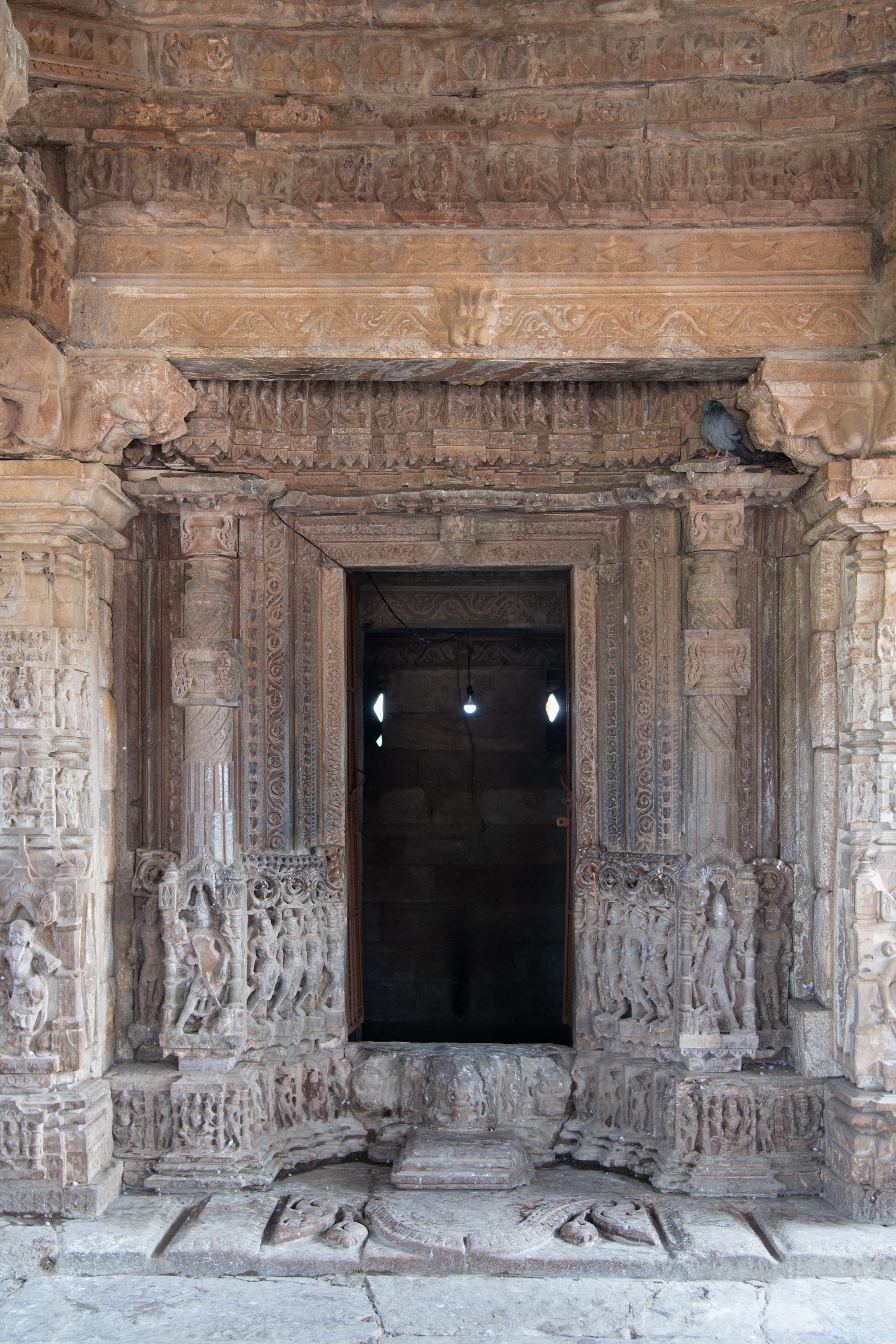 The mandapa (pillared hall) leads to the antarala (vestibule or antechamber), a shallow passage distinguished by pillars along its sides. From there, one proceeds to the garbhagriha (sanctum sanctorum) of the Undeshwar Temple, featuring a meticulously carved doorframe. This doorframe exhibits a multi-shakha (vertical band or architrave) design, with each shakha adorned with various floral patterns, naga (serpant) figures and stambha (buttress). At the lower part of the doorframe, there are four female figures, two of whom hold kalashas (pitchers) while the other two hold chauris (fly whisks). Following them are four-armed dvarapalas (door guardians), each depicted with their respective attributes. Lastly, there is a figure of Kubera holding a money bag. Above the lalatabimba (lintels) on the doorframe's entablature, Nataraja is positioned at the centre, flanked by seated goddesses, with Brahma and Vishnu depicted at the ends.