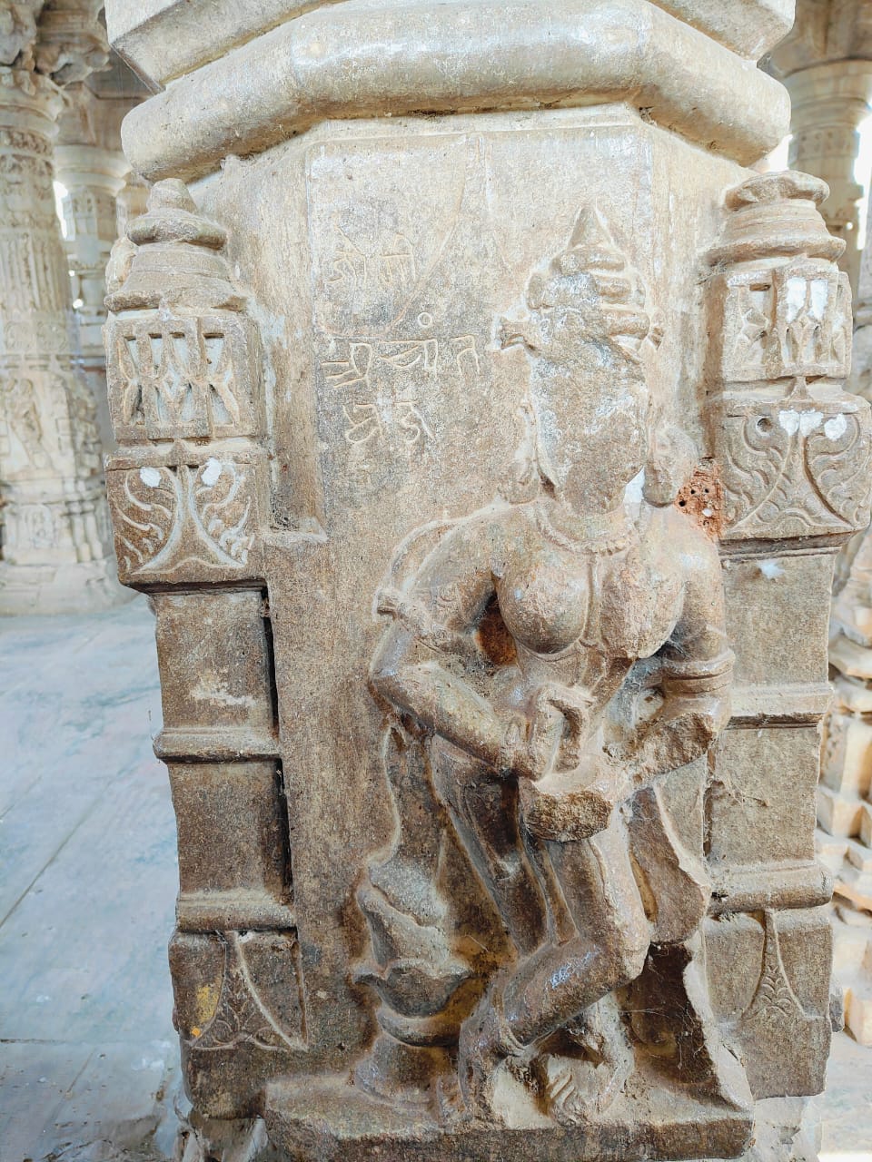 Unlike other apsaras (celestial damsels) carved on the shaft of pillars, this particular figure is unique. She is crowned and holds a cup in her left hand while her right hand holds something dipped in the cup. Similar images from the Harshnath Temple Complex depict a cup being held, symbolizing the nectar of bliss attained after the completion of tantric sadhana, while the fingers of the other hands are sometimes dipped into the cup. However, connecting this obscure image of a damsel with tantric connotations might lack substantial support.