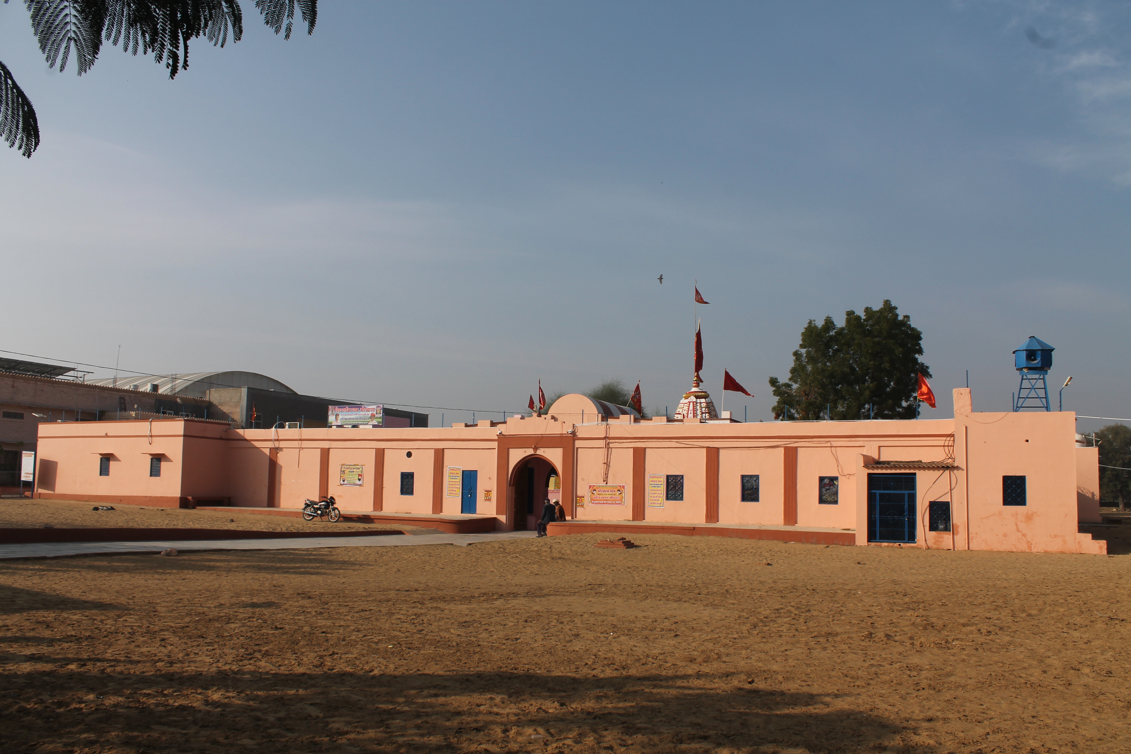 View of the modern dharmashala (a charitable public rest house or shelter) rooms added surrounding the verandah (open-air hallway or porch) of the Suswani Mata temple complex. The shikhara (superstructure) of the main temple and the flagstaff of the centrally located old temple are visible from the open ground in front of the main gate.