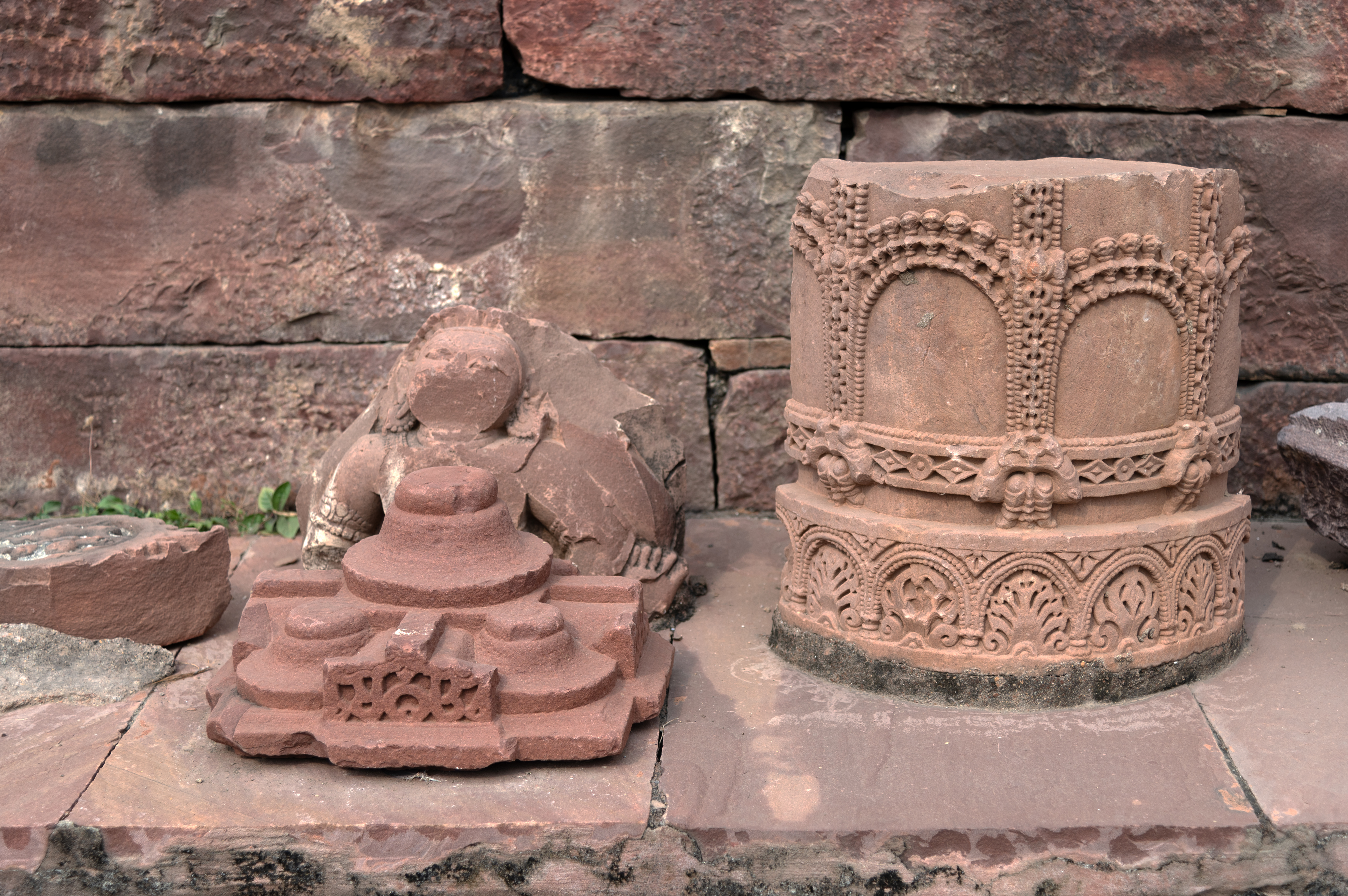 The Mahanaleshwar temple complex's premises host various architectural fragments scattered next to the pratoli (entrance gateway). Seen here are fragments of a pillar capital, part of the samvarna shikhara (pyramidical superstructure) of the mandapa (pillared hall), and a pillar fragment. The round shaft remnant of the pillar bears minute carvings.