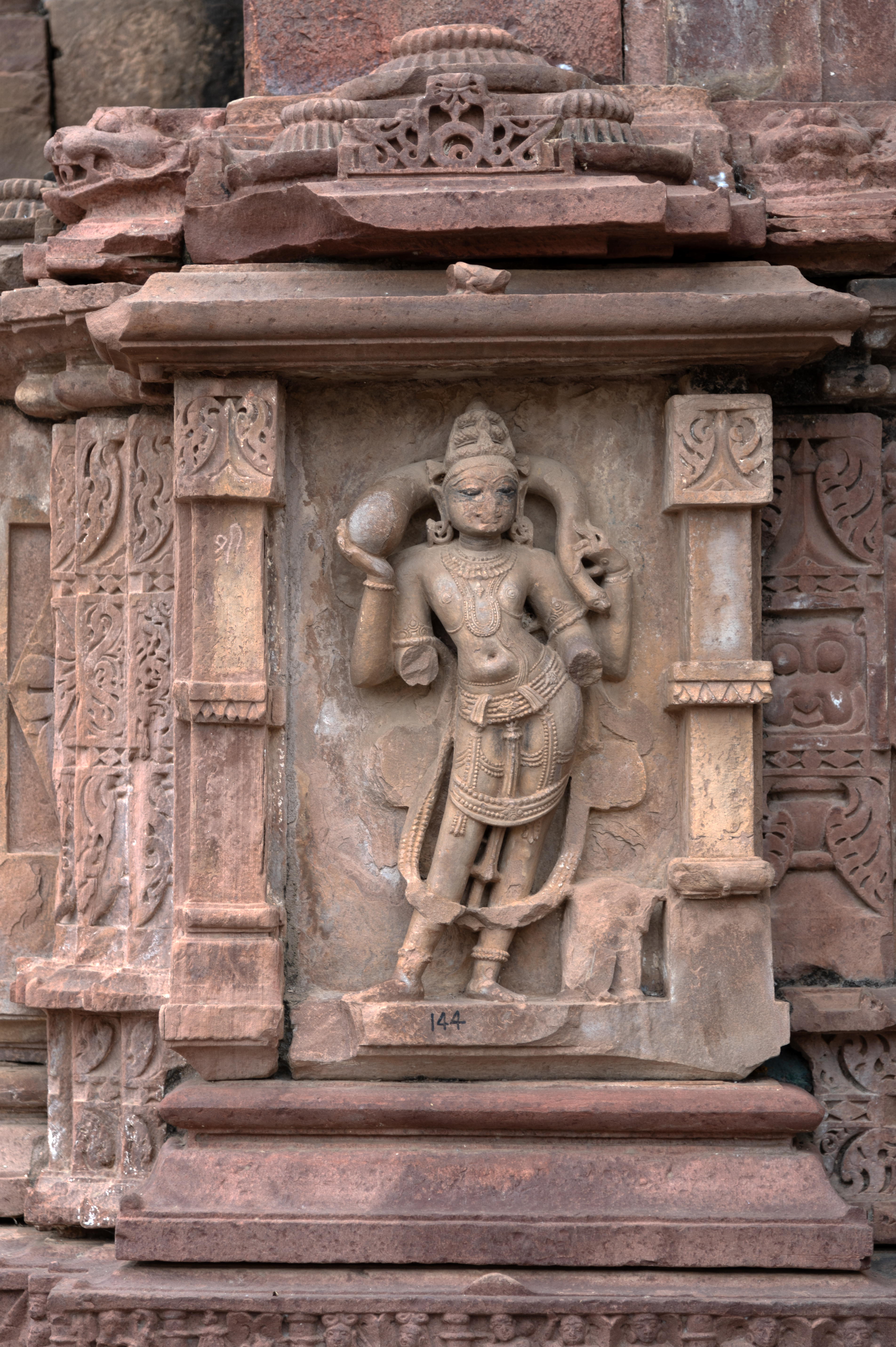 The Mahanaleshwar Temple's dwarf walls offer a close-up view of Kubera, one of the dikapala (gods of the cardinal directions), in the devakoshtha (niche). Kubera corresponds to the northern direction. One of Kubera's distinctive attributes is the broken lower two hands of the deity, while the upper two hands hold a nakula (money) bag. A miniature samvarna (pyramid-shaped) variety of shikhara (superstructure) surmounts two slender pilasters to form the devakoshtha. A udgama (gavaksha, or dormer window design) fronts the ghantas (bells) of the shikhara. It rests on a low pedestal made up of multiple mouldings.