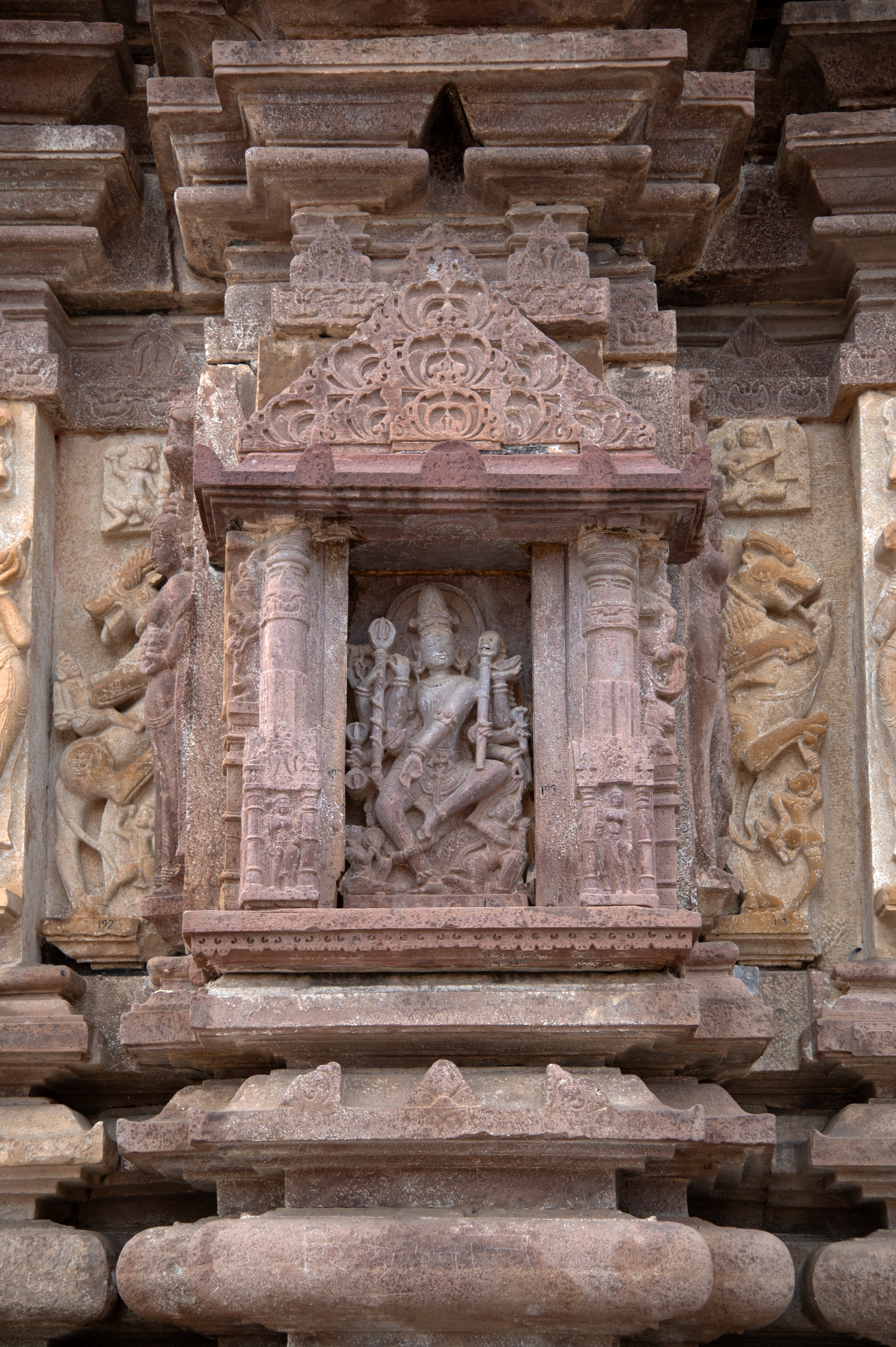 The central bhadra of the east-facing jangha of the temple has a Natesha icon. The devakoshtha that frames the Natesha is similar in style to the one on the east-facing jangha. The eight-handed Natesha is depicted in a dancing pose, holding a snake, kapala, khatvanga, trishula, and damaru attributes. Natesha has a jata mukuta (matted hair) and a halo behind his head. At Natesha's feet, there are two dancing figures who are likely attendants.