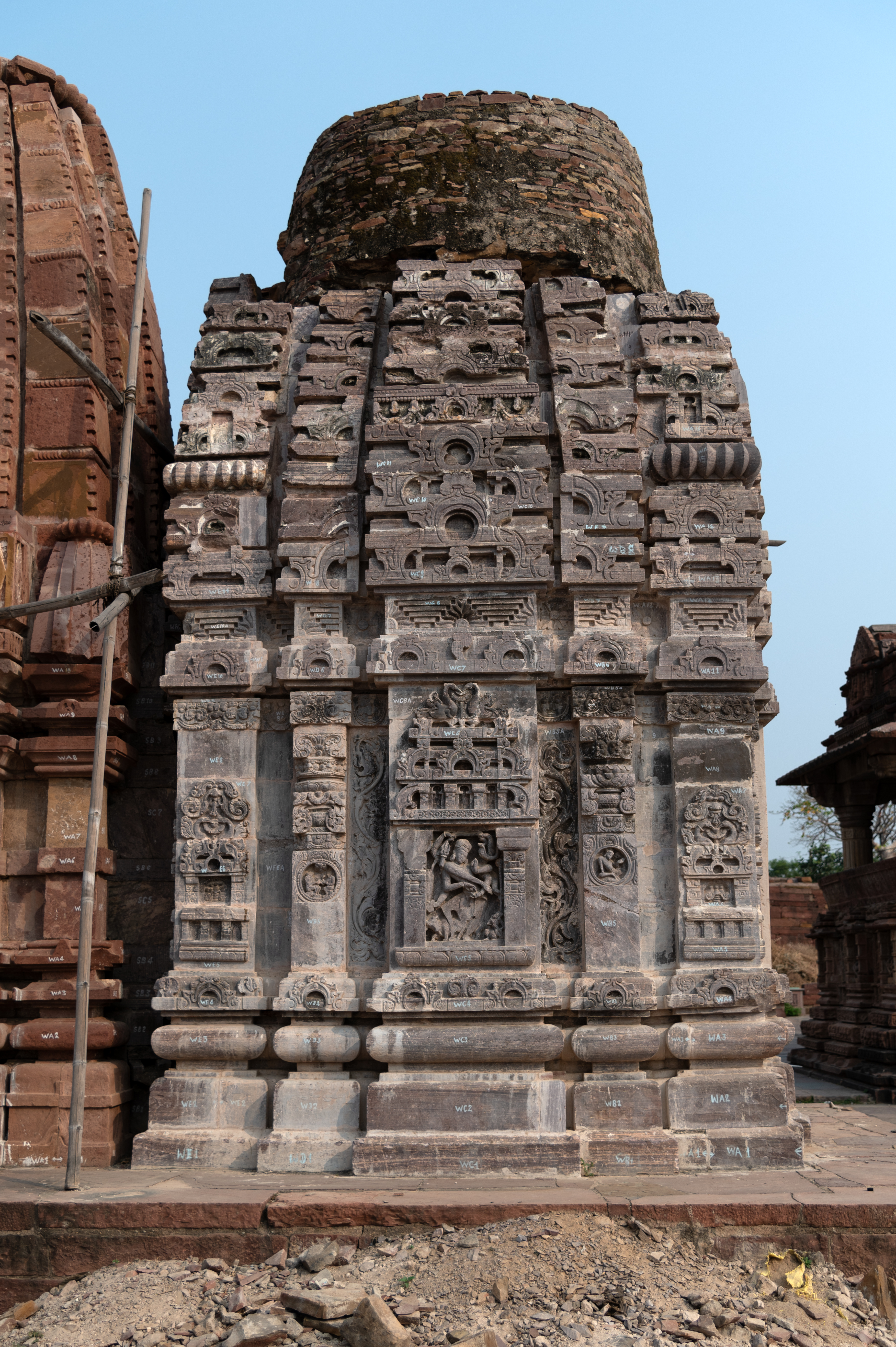 This is the west-facing back elevation of the southern shrine from the Triple Shrine Temple in the Mahanaleshwar temple cluster. This is a single-shrine temple built in the latina (mono-spire) style of temple architecture. Only two levels of the shikhara (superstructure) are in situ. The vicinity contains other architectural fragments of the temple. Except for the bhadra projection, the temple does not have any sculptures on the exterior wall surfaces. The ornamentation of the temple mainly consists of geometric patterns, floral motifs, and architectural decorations.