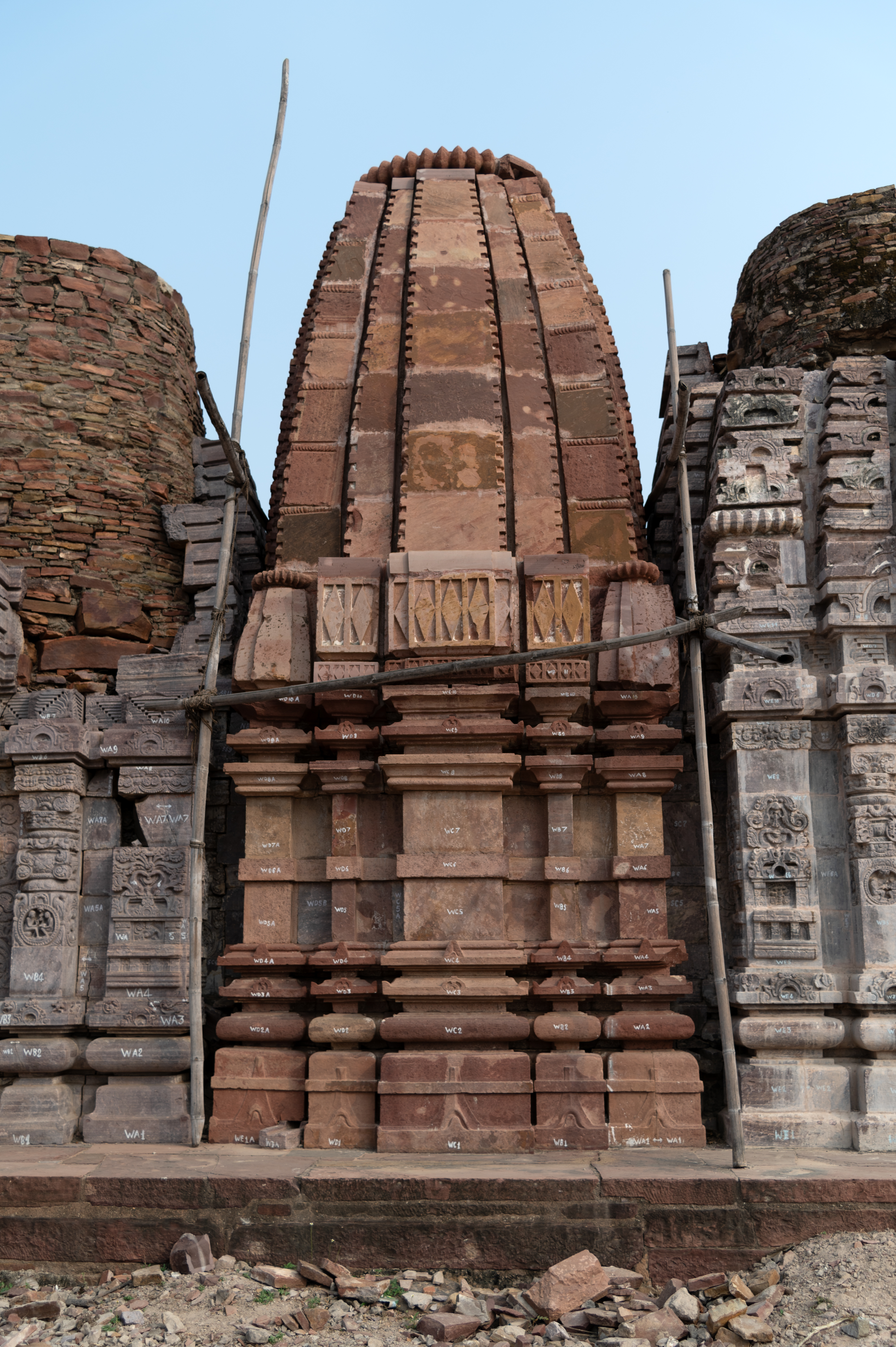 The Triple Shrine's central shrine is a later addition to the edifice. Built in red sandstone, this single-shrine temple is also adorned with a shikhara (superstructure) of the latina (mono-spire) variety. This shrine's base mouldings and wall feature a pancharatha design, which consists of five divisions to the plan and elevation. The shikhara's finial has an amalaka (ribbed disc). The temple exterior does not have any sculptures or ornamentation. The latina shikhara has small aedicules on the corners and a mulashringa (main spire) consisting of five bands. The shikhara's corner bands are made up of seven blocks separated by flat ribbed discs.