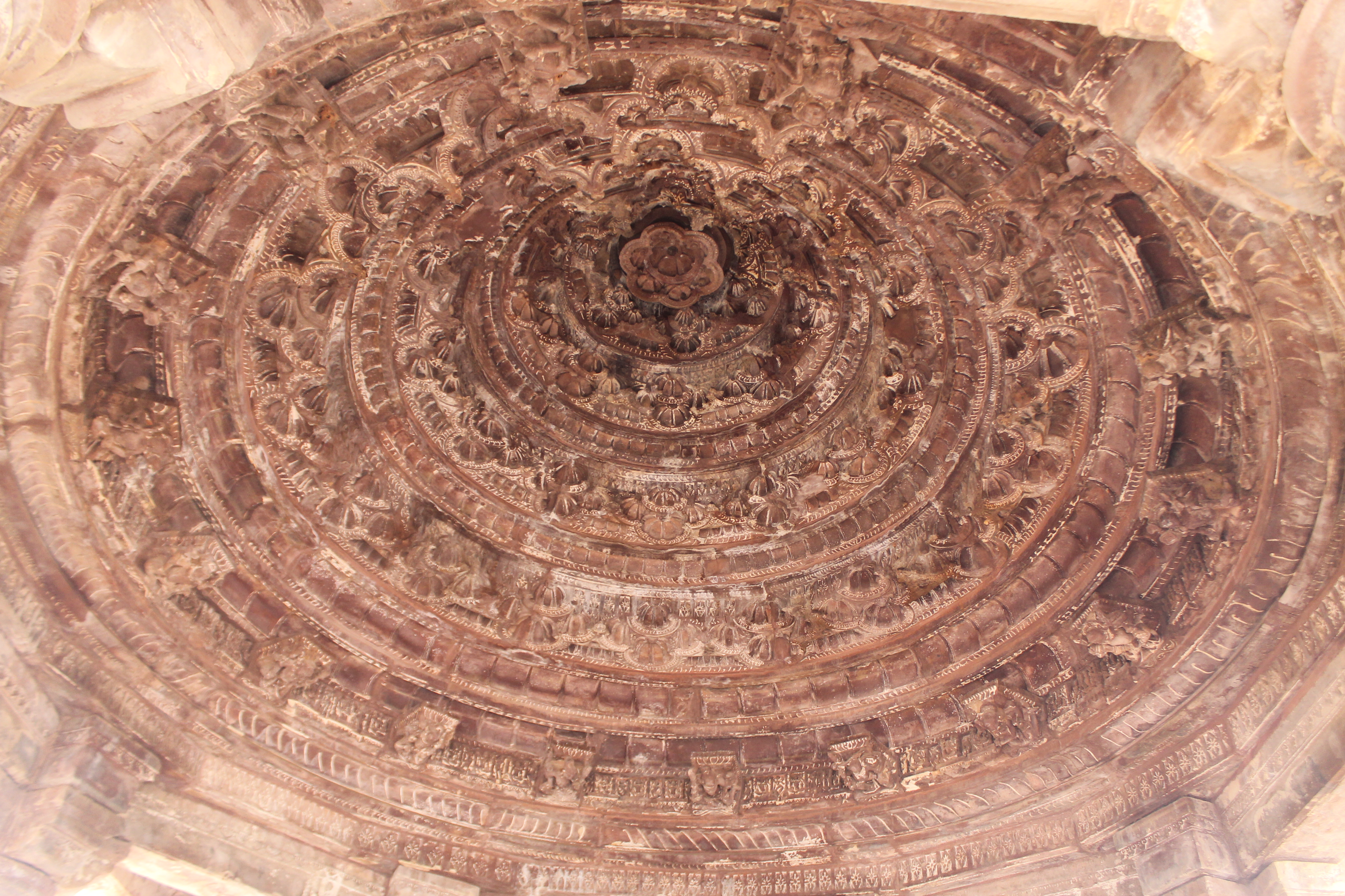 The domical ceiling in the Suhaveshwar Temple's mandapa (pillared hall) is seen here. The ceiling consists of concentric circles composed of small cusps. Each inner circle has a slight offset from the outer circle. The ceiling's center is adorned with a suspended lotus medallion. The ceiling rests on eight pillars. Just above the pillar capitals, the circular portion of the ceiling has small square brackets with sculptures.