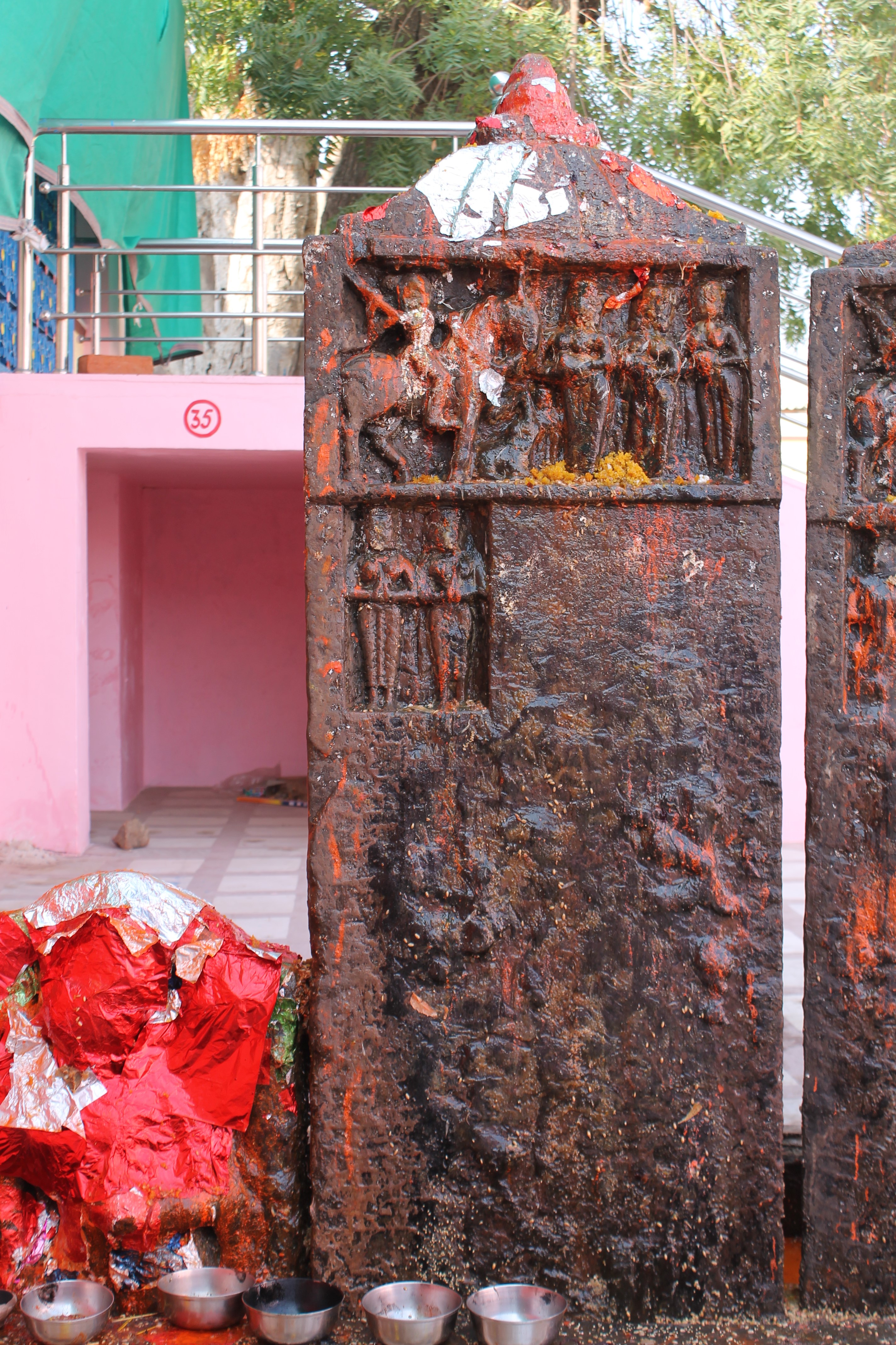 Image 3: Seen here is an inscribed and sculpted hero stone (memorial stone) in the Suswani Mata temple premises, which is built in memory of the deceased soul. It is approximately five feet in height, and the front face has an inscription at the centre and human figures in the upper part. The stele is capped with a shikhara (superstructure or spire) motif.