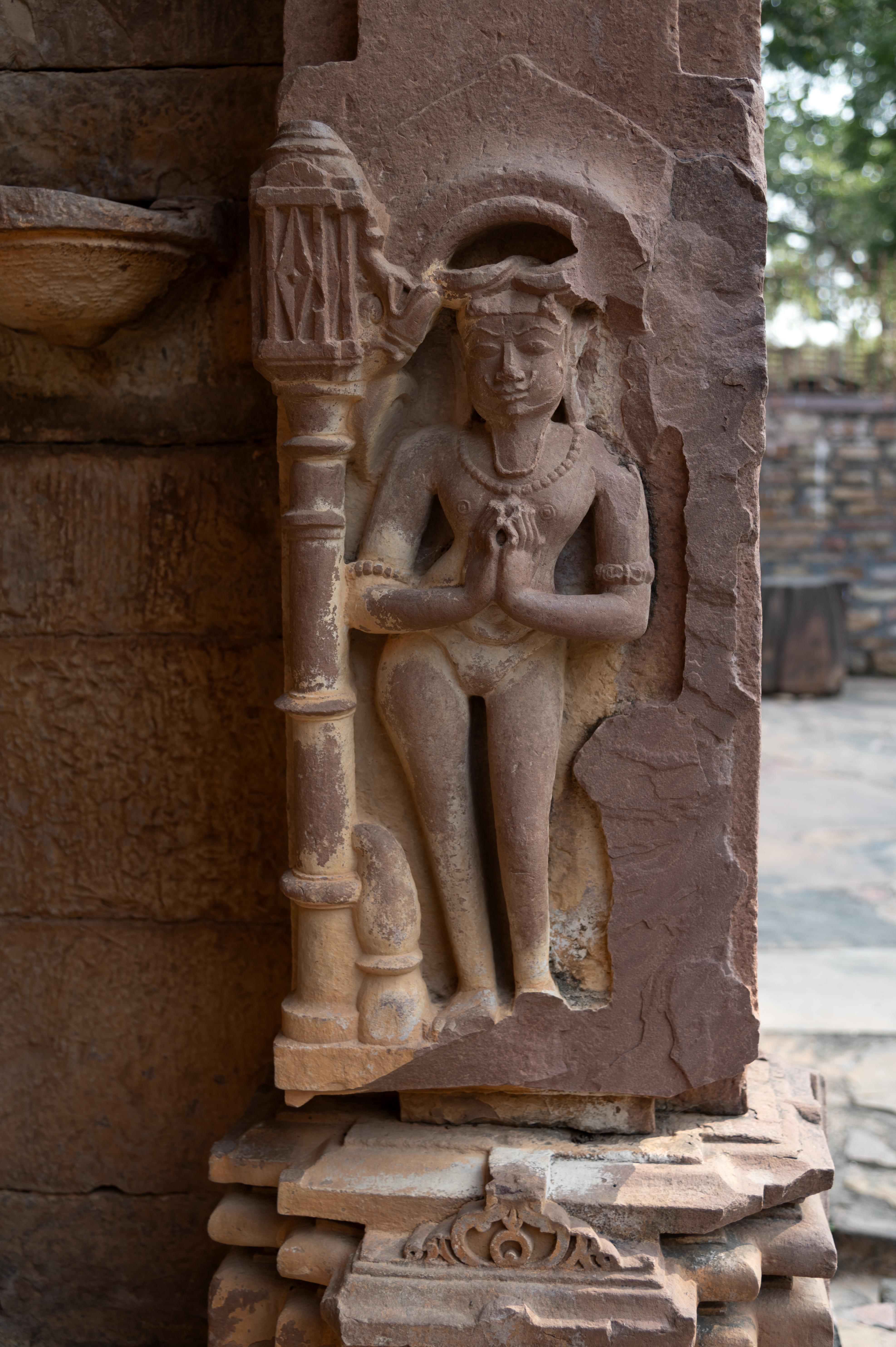 The pilaster on the left side of the antarala (vestibule or antechamber) of the Kaner-ki-Putli Temple depicts a standing ascetic with folded hands. He has thick, matted hair locks, a long beard, and a moustache. His upper body is bare, while his lower body is clad in a langot (loincloth).