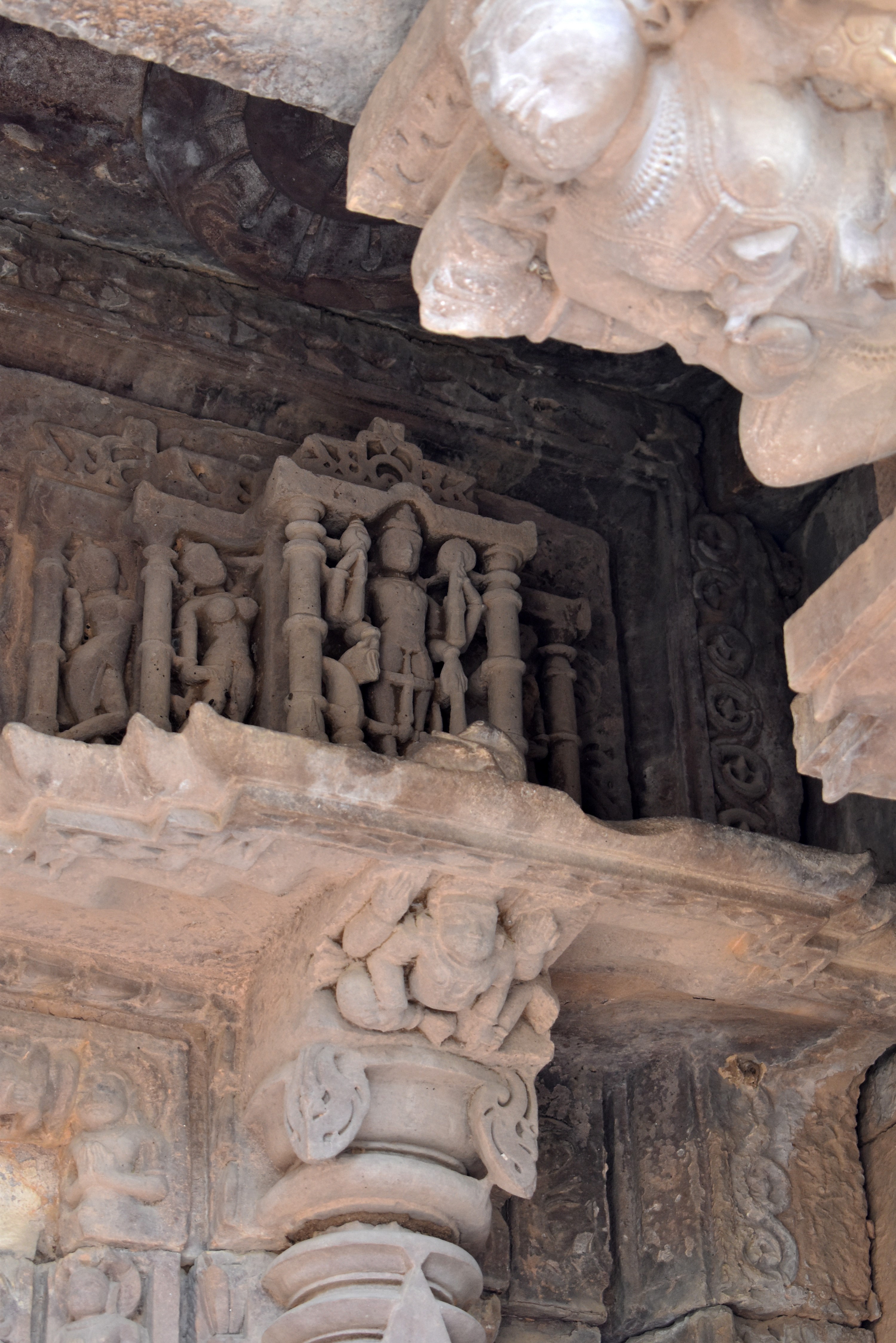 Vishnu is positioned at the far left end of the uttaranga (entablature) above the lintel of the garbhagriha (sanctum sanctorum). He is depicted holding a gada (mace), a shankha (conch shell), and a chakra (discus). One of his lower hands is in the varada hasta (boon-giving gesture). He is adorned with a kiritamukuta (a cylindrical crown associated with Vaishnava iconography) and various pieces of jewellery.