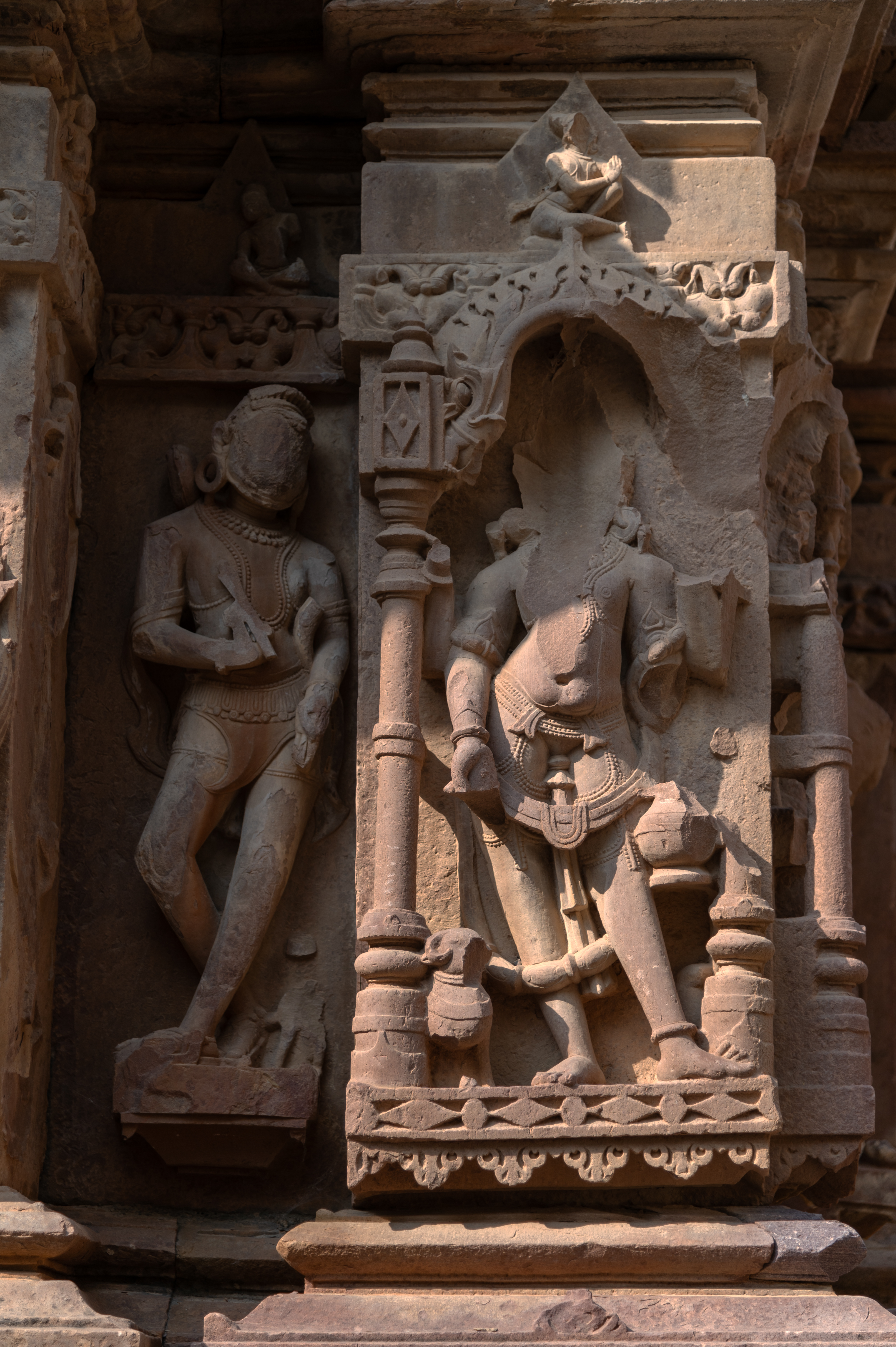 In a niche on the southern wall of the garbhagriha (sanctum sanctorum) of the Kaner-ki-Putli Temple, a male figure is depicted. Unfortunately, the figure is damaged, with the head entirely missing. Both upper arms are broken, while the figure holds a kamandalu (water carrier or pot) in the left hand and makes a varada hasta (boon-giving gesture) with the right hand. At the base of the niche, a broken ram-like animal with horns is visible. Based on the vahana (mount), attributes, and gestures, this figure can be recognized as Agni (one of the deities of cardinal directions). Adjacent to Agni, there is a male figure that stands out from the others. This figure features curly, raised hair and a beard, but unfortunately, the face has been eroded. The upper body is bare and adorned with jewellery, while the lower body is covered by a distinctive triangular loin cloth. An antaravastra (inner garment) is visible beneath the loin cloth. The figure is draped with a scarf wrapped around the arms, and although the broken object held in his two hands is now incomplete, the visible middle part resembles a stick. Notably, this figure is unique because of the distinctive dressing style and footwear, suggesting him to be a foreigner.