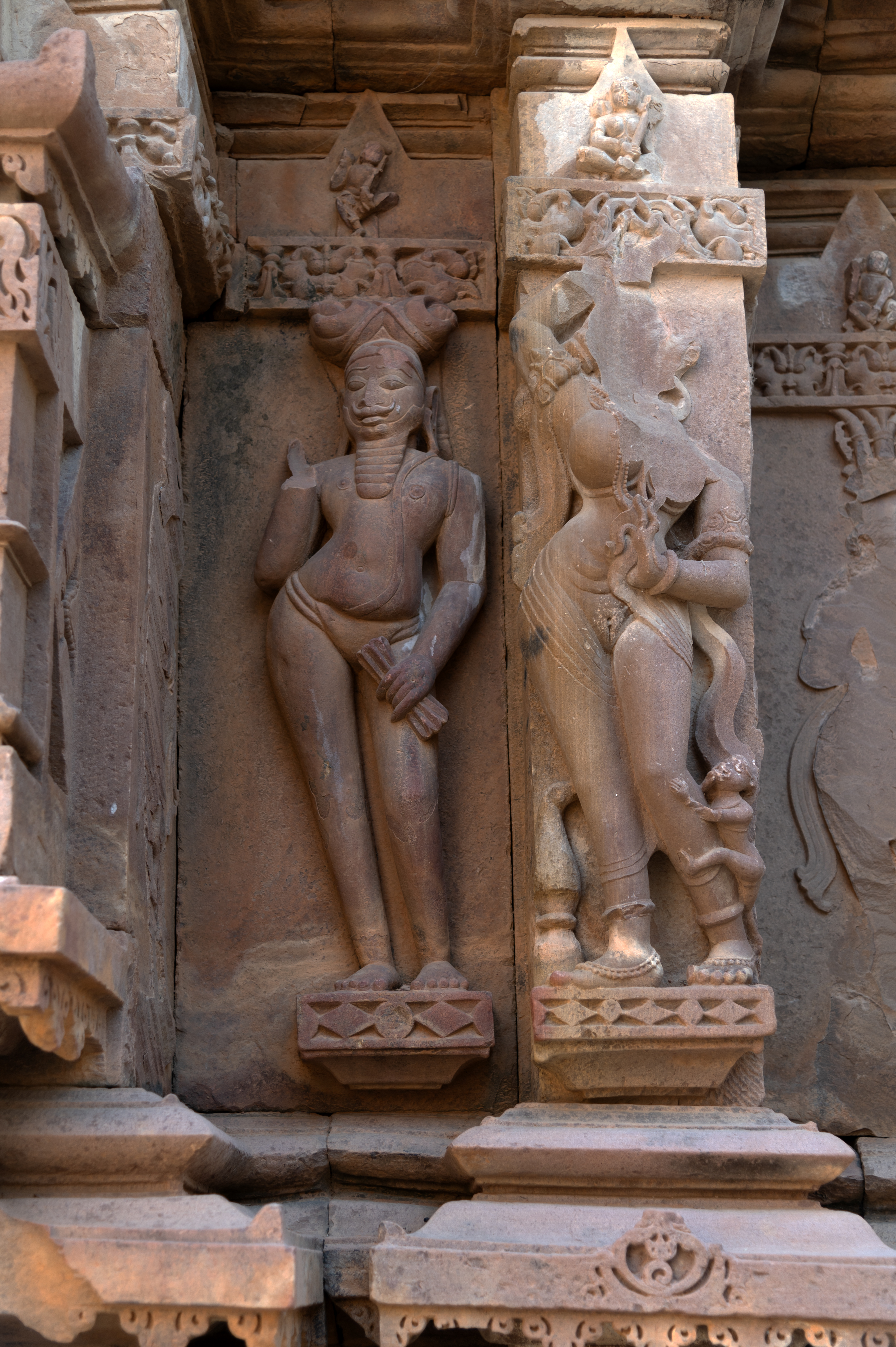 The details of the rear or eastern wall of the garbhagriha (sanctum sanctorum) illustrate the contrast between the ascetic and the surasundari (celestial damsel). The ascetic (on the left) is characterized by a substantial jatabhara (braided hairdo) atop his head, a lengthy beard, and a moustache. His elongated earlobes signify the mahapurusha lakshana (markers of a great individual). His bare upper body is adorned with a broad yajnopavita (sacred thread), with a langot (loincloth) covering his lower body. Despite the apparent damage and raised position of his right hand, his left-hand holds an unidentified object. The damaged surasundari figure, on his right, stands gracefully with a voluptuous body adorned with exquisite ornaments. Though her raised right hand is damaged, she holds a fine scarf in her left hand near her belly, draping it around her arms. Her lower body is covered by a translucent garment so delicate that her body underneath is visible. A small creature is depicted climbing her left leg. In stark contrast to the tranquil and composed posture of the ascetic, the surasundari vividly embodies sensual beauty. Notably, there is a kalasha (pitcher) placed at the bottom near her feet, surrounded by foliage symbolizing abundance and growth, mirroring the symbolism of the female body.