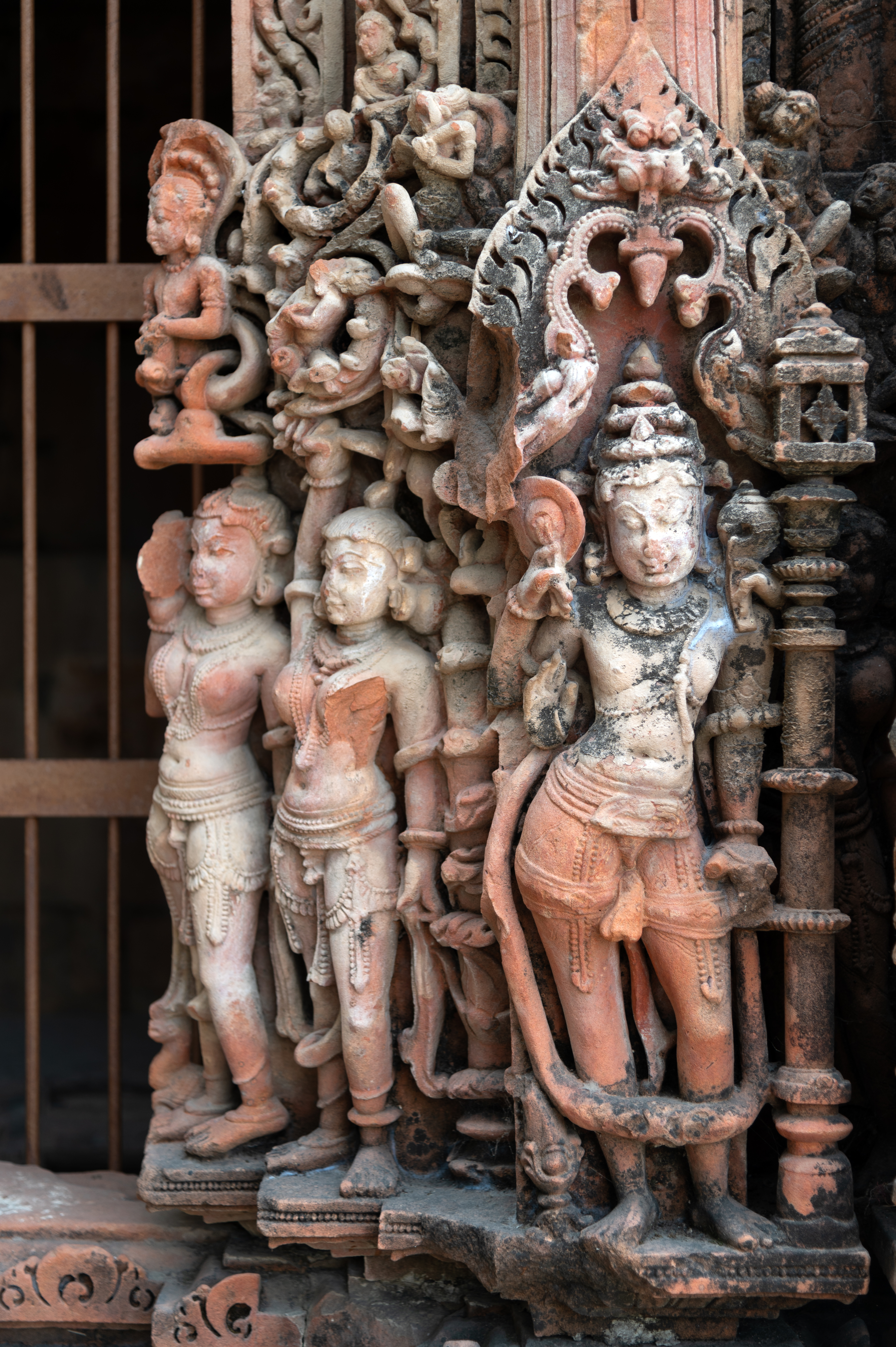 The lower section of the garbhagriha (sanctum sanctorum) entrance to the Vishnu Temple displays the river goddesses, Ganga and Yamuna, followed by a chauri (fly whisk) bearer, and then the Vaishnava dvarapalas (door guardians). The depiction shows these four-armed dvarapalas holding a gada (mace), a shankha (conch shell), and a chakra (discus), with one hand in the abhayahasta (a hand pose indicating reassurance or protection from fear) position. A karand mukuta (tiered crown), long garlands, and elaborate jewellery adorn them.