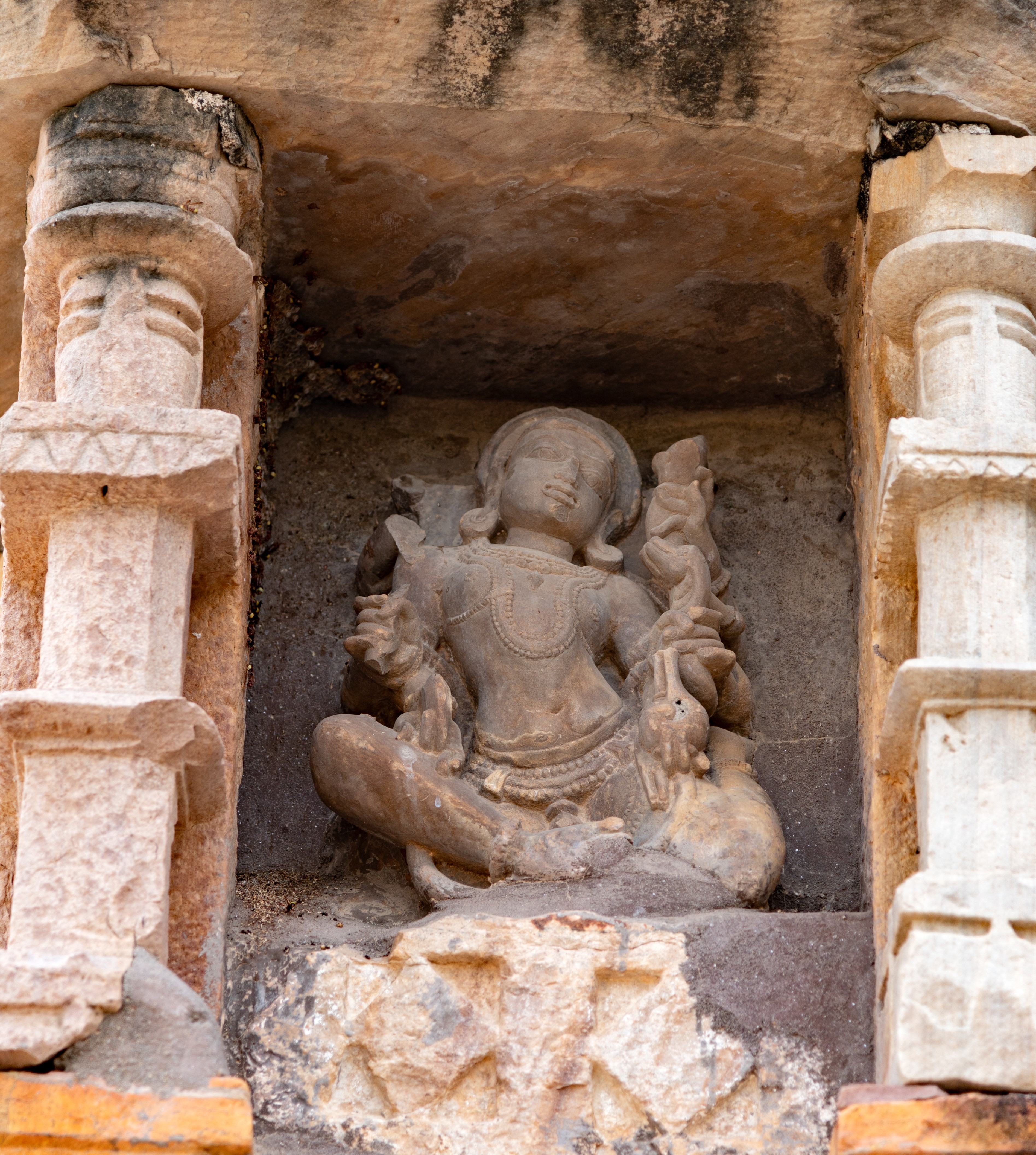 The figure installed in the bhadra (central offset) niche of the lateral wall of the garbhagriha (sanctum sanctorum) is particularly noteworthy. This unidentified deity is depicted as moving through the air, with one leg raised backwards. Although many of the deity’s multiple arms are broken, the intact hands display a shankha (conch shell) and a chakra (discus) in the front right and left hands, an unidentified object in the middle left hand, and a pushpa (flower) in the upper right hand.