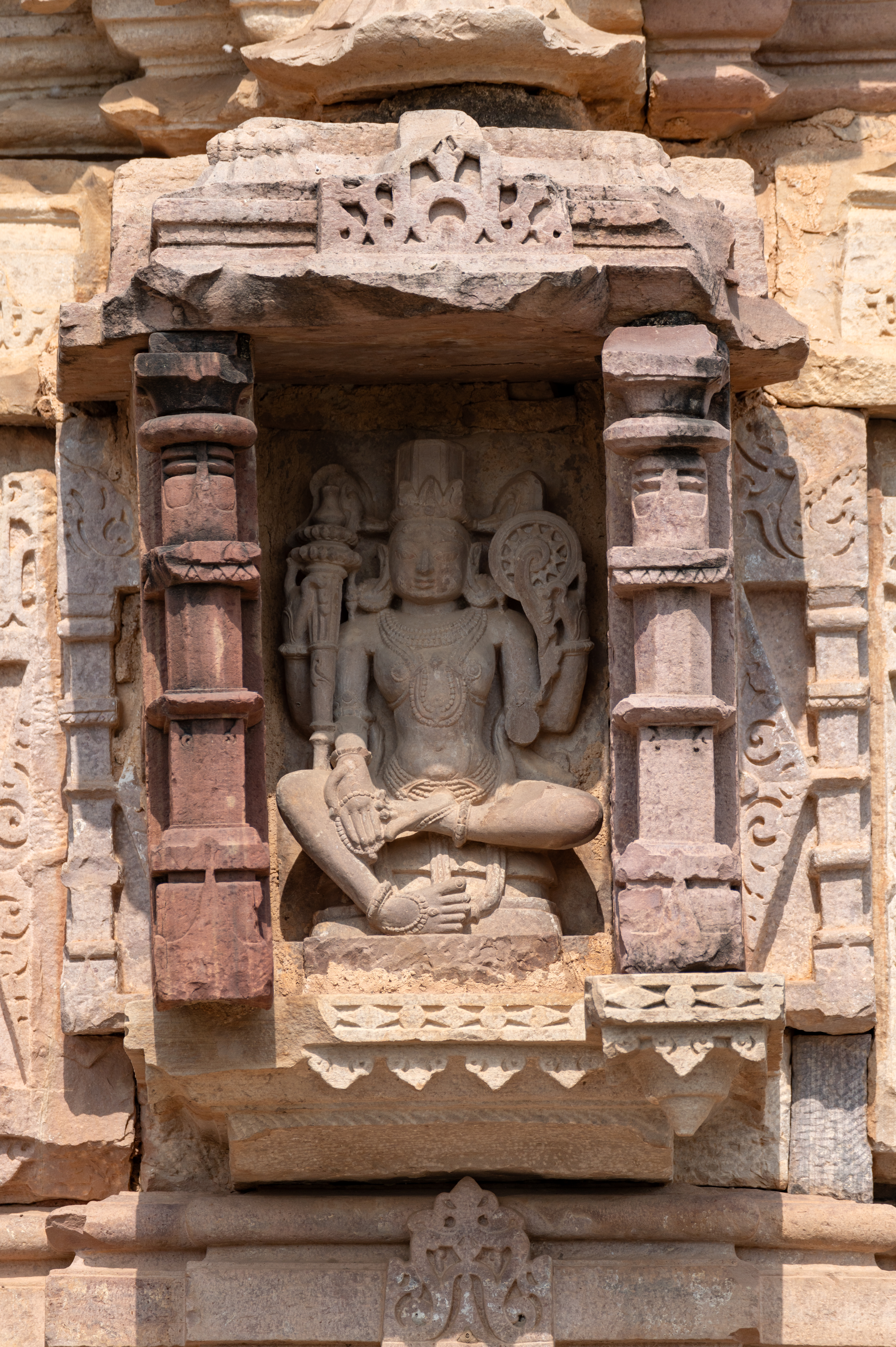 The bhadraratha (central projection) features a seated Vishnu, intricately carved with four arms. Crowned, Vishnu holds a gada (mace) and a chakra (discus) in his upper hands. His lower right hand holds an akshamala (rosary) while also making a varada hasta (boon-giving gesture). Although his lower left hand is broken, Vishnu is adorned with large circular earrings, a broad beaded necklace, a breast band, a girdle, bangles, and anklets.