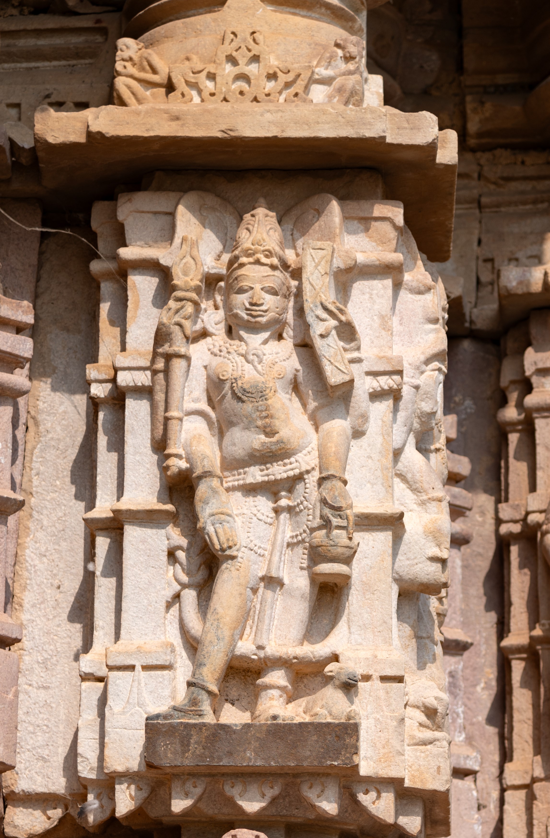 The four-armed Agni is depicted with a shruka (sacrificial ladle) and a pustaka (book) in his upper hands. His lower right hand makes a varada hasta (boon-giving gesture), while his lower left hand holds a kamandalu (water pot). He is adorned with a jatamukuta (crown of matted hair), a moustache, and a long, intricately knotted beard. He is bejewelled and accompanied by his mount, the ram.