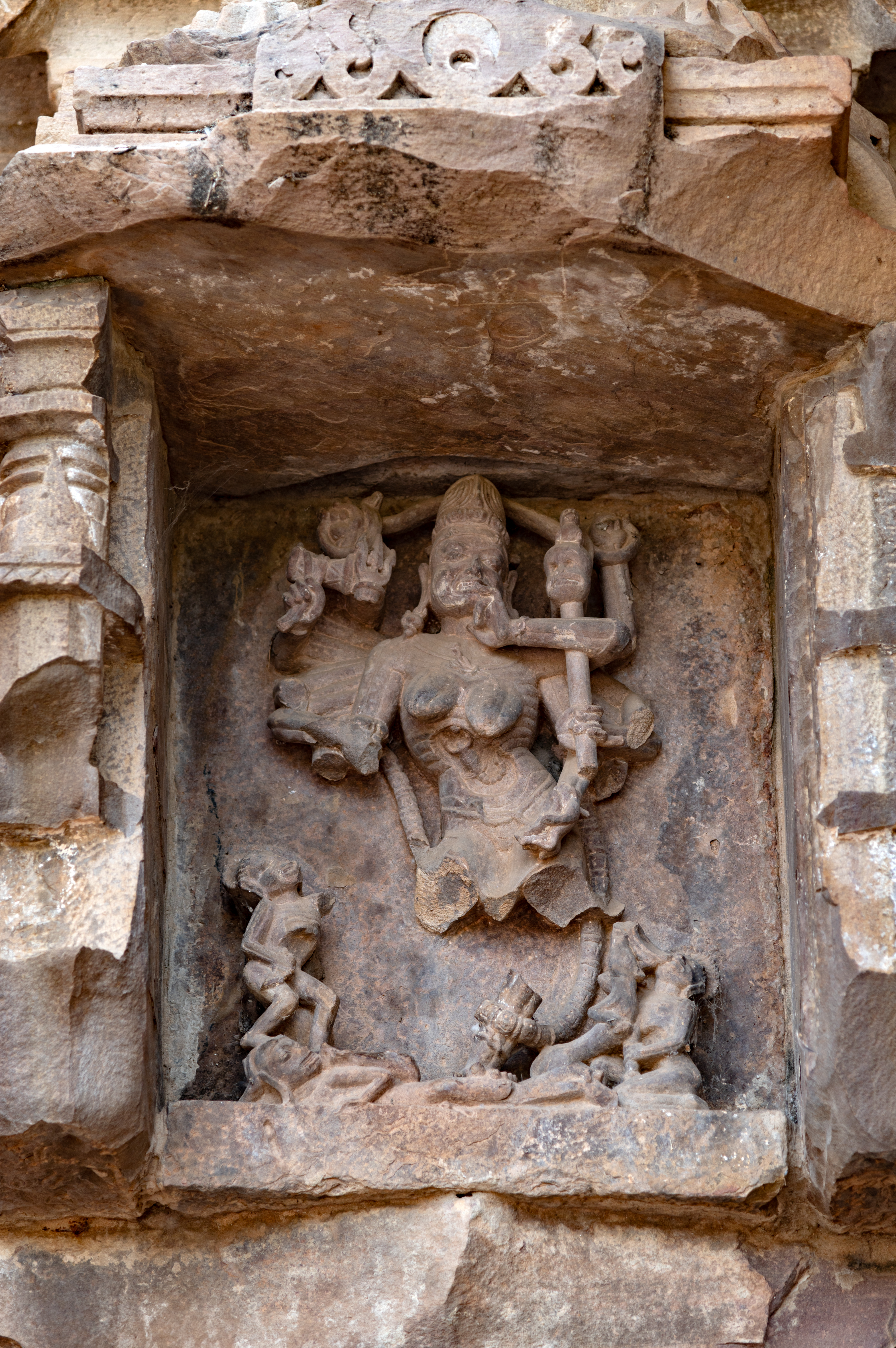 Chamunda stands atop a human form, flanked by two attendants and an animal companion. Though many of her arms are fragmented, her divine attributes are evident. In her upper hands, she carries a sarpa (snake). Her intact right arms carry a damru (drum) and a trishula (trident), while her left hands cradle a khatvanga (staff with a human skull) and a bowl, with the finger of her left hand touching her lips. Chamunda’s portrayal emphasizes her skeletal form, with a cavity in her belly housing a scorpion, symbolizing transformative power. Her lower body is draped modestly with a loin cloth. Adjacent to her, a seated figure grasps a bowl from which he is drinking. It is speculated that one of the goddess’s broken hands may have held a munda (severed human head). The presence of the seated figure suggests the consumption of the blood dripping from the severed head.