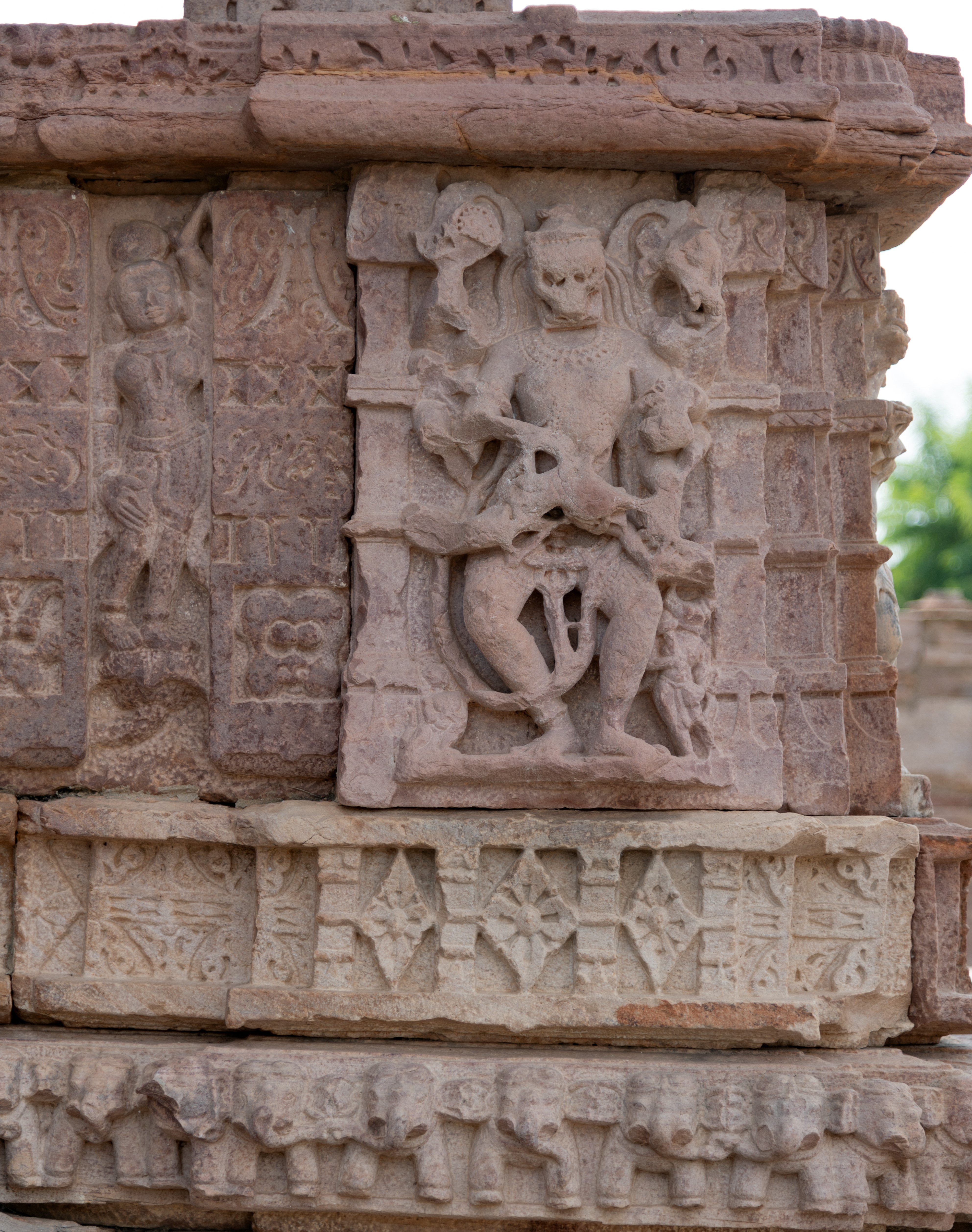 Narasimha is carved on the external wall of the mandapa (pillared hall), though time has weathered its intricate details. This depiction portrays the fourth incarnation of Vishnu, showcasing his formidable aspect with a lion’s visage atop a human form. Despite the erosion of many arms, Narasimha’s attributes are intact. In his upper hands, he holds a chakra (discus) and a shankha (conch shell), symbols of cosmic order and divine sound. His frontal hands are depicted in the act of subduing an asura (demon), although the figure is heavily damaged. He is adorned with jewels and a long garland. Additionally, a figure resembling Varaha is carved at the bottom left.