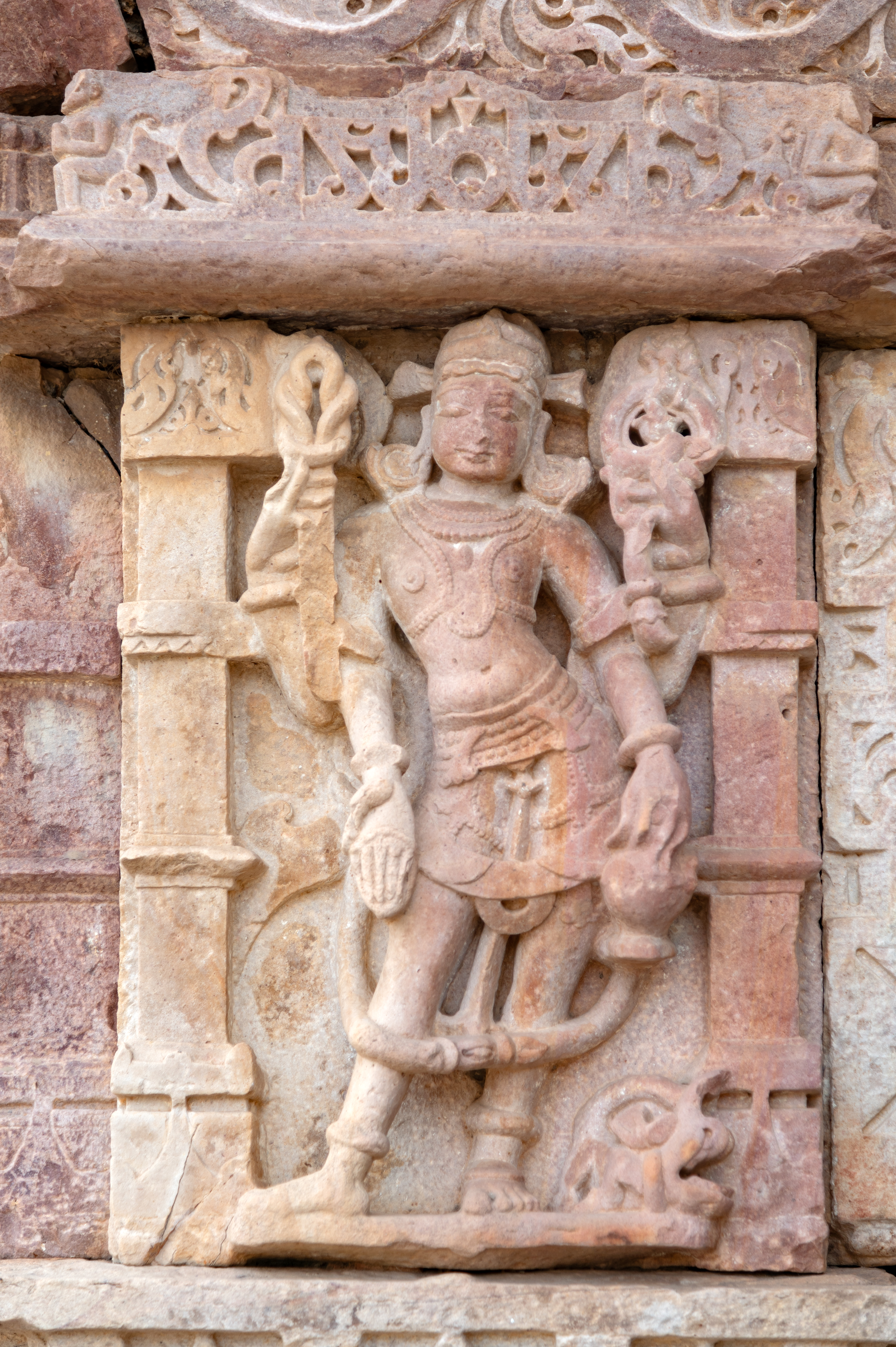 Varuna is depicted alongside his vahana (mount), the makara (crocodile-like mythological sea creature). In his upper right hand, he holds a pasha (noose), symbolizing his control over the cosmic forces. In his upper left hand, he holds a pushpa (flower), representing his connection to the natural world. His lower left hand holds a kamandalu (water pot), signifying purity and sustenance, while his lower right hand extends in varada hasta (boon-giving gesture).