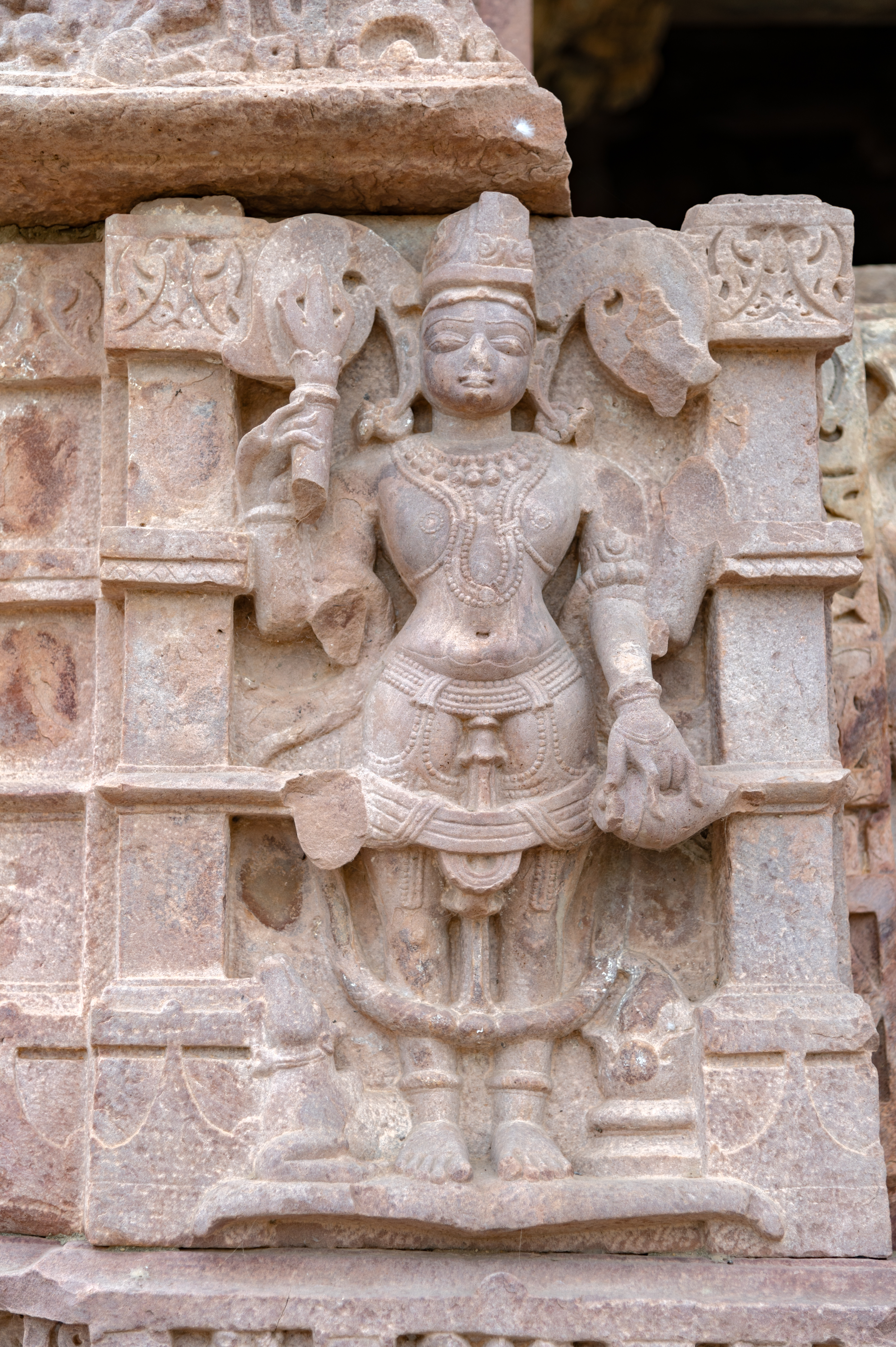 Harihara is depicted as a synthesis of Shiva and Vishnu. In this form, Shiva occupies the right half, adorned with a jatamukuta (crown of matted hair). Vishnu occupies the left half, crowned with a kiritamukuta (a cylindrical crown associated with Vaishnava iconography). In his upper right hand, Harihara wields a trishula (trident), symbolizing Shiva’s power, while his lower left arm holds a shankha (conch), symbolizing Vishnu’s presence. Nandi, Shiva’s mount, is carved at the bottom right, and a kalasha (pitcher) adorned with vegetal motifs is depicted at the bottom left, symbolizing abundance.