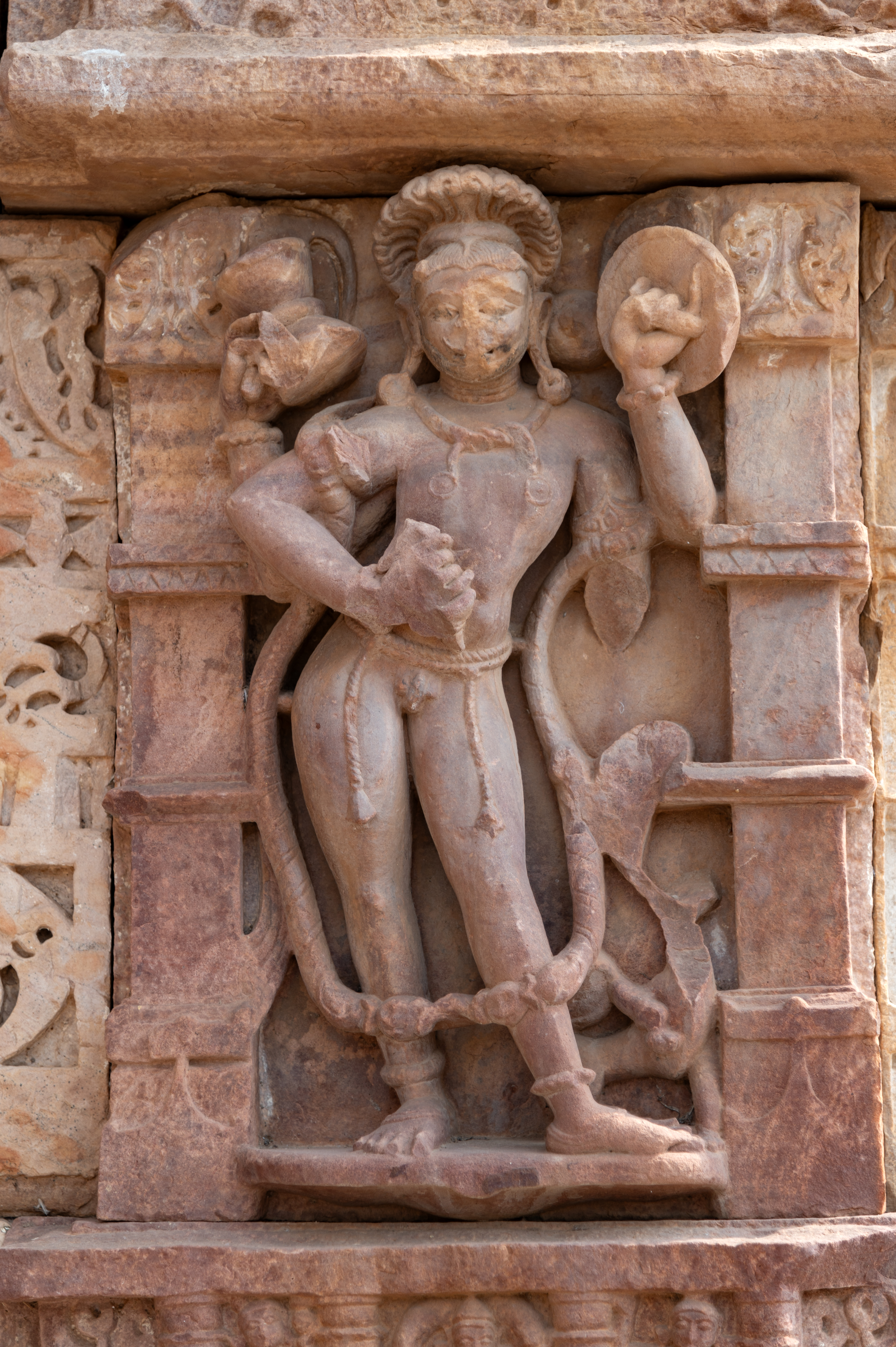 A figure of Nirutti stands prominently with a noticeable phallus, symbolizing his association with fertility and vitality. In his hands, Nirutti holds a damru (drum), a khetaka (shield), and a musala (pestle). At the bottom left, a broken animal is depicted. Nirutti’s distinctive high-raised hairdo adds to his unique presence, while his adorned appearance with jewels and a long garland underscores his divine status.